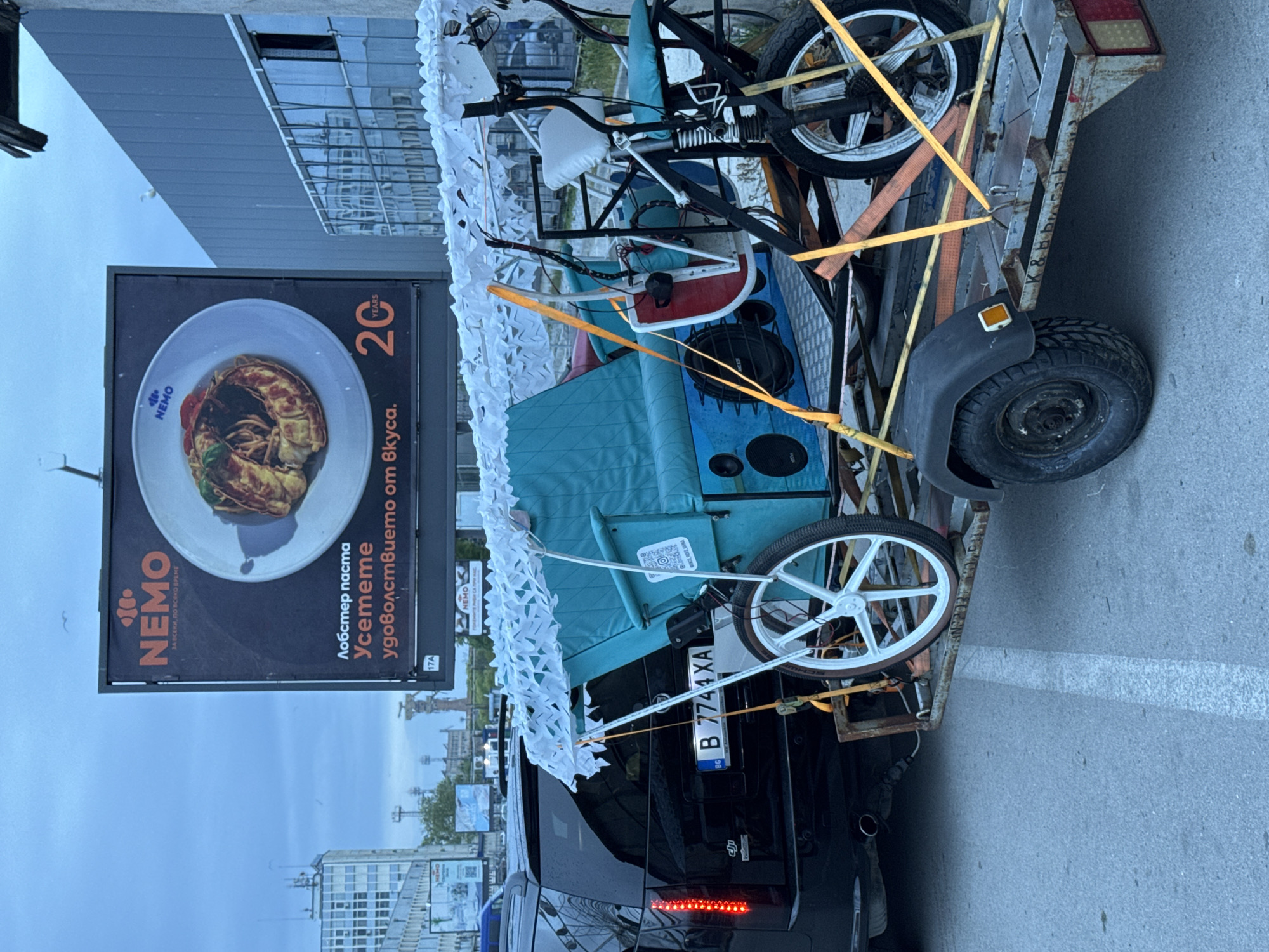 The image shows a black SUV with a trailer attached to it, parked near a large billboard in Varna, Bulgaria. The billboard advertises a restaurant called Nemo, featuring an image of lobster pasta and the text "Лобстер паста Усетете удоволствието om Bkyca." (Lobster pasta. Feel the pleasure of the taste.) and "ЗА ВСЕКИ, ПО ВСЯКО ВРЕМЕ" (For everyone, every time). The billboard also indicates the location of Nemo as 17A. The license plate of the SUV is B 7744 XA.
The trailer is loaded with a variety of items, including a bicycle and an aqua-colored vehicle with speakers. These items are secured with yellow straps. A white net-like material is draped over part of the vehicle on the trailer. The background features urban buildings and a cloudy sky, suggesting an overcast day in Varna.