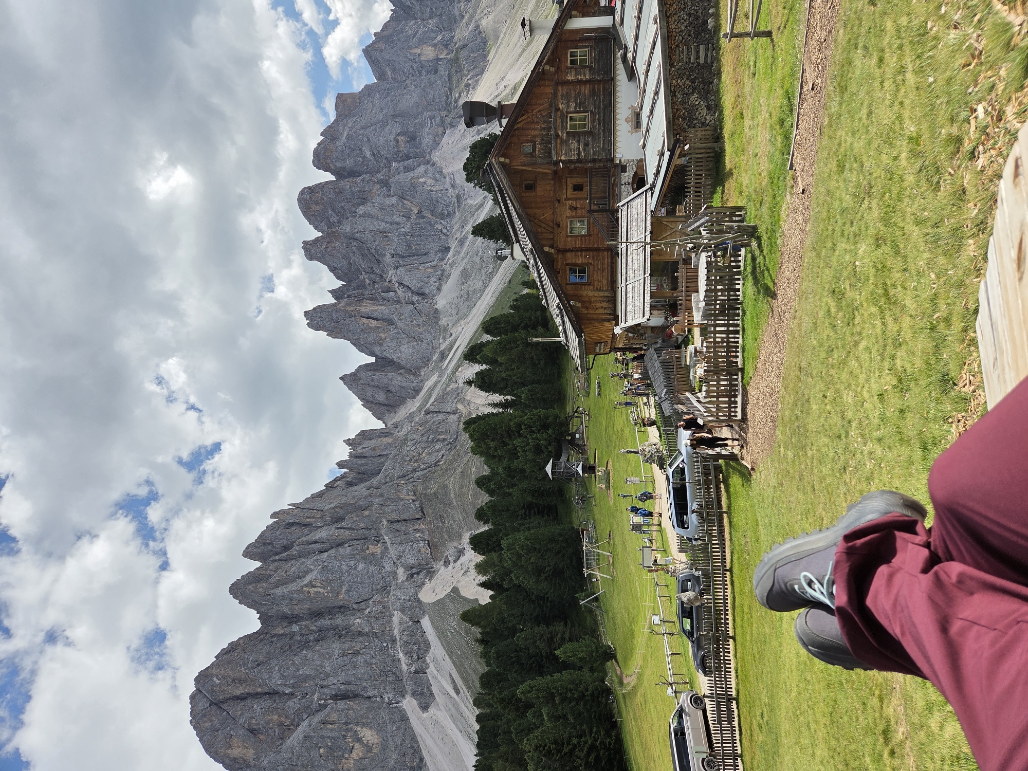 In an outdoor scene, a person's legs and feet are visible in the foreground, resting on a wooden surface. The person is wearing burgundy pants and dark grey athletic shoes with light blue laces. The background features a grassy hill with a wooden fence and scattered picnic tables. Beyond the fence, several vehicles are parked on a gravel area. Further back, a traditional wooden mountain hut with a dark roof and a chimney stands prominently. A dense forest of evergreen trees lines the base of a towering, jagged mountain range, whose rocky peaks are partially obscured by a cloudy sky. Several people are walking and standing near the mountain hut and on the grassy area. The overall scene depicts a serene natural landscape with a rustic dwelling.