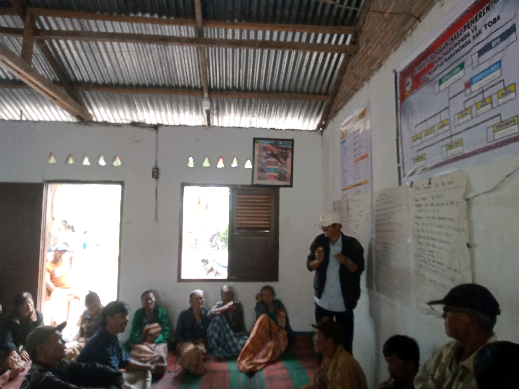 The image depicts an indoor gathering of approximately 15 people in what appears to be a simple, rustic building. The room has a corrugated metal roof supported by wooden beams. The walls are white, with several small, triangular openings for ventilation. A framed picture hangs on one wall, and a large organizational chart is displayed on another. Natural light enters through doorways and windows.
In the foreground and midground, a group of individuals are seated on the floor, some on mats. The individuals are diverse in age and gender, dressed in casual attire. Some are looking towards a man standing in the center of the room, who is wearing a white cap and a dark jacket over a blue shirt. He appears to be addressing the group. To the right of the standing man, another man is visible, wearing a dark cap and a light-colored shirt. In the background, through an open doorway, more people and some activity can be seen. The setting suggests a community meeting or information session, possibly within Narumonda 6, Indonesia, given the presence of an organizational chart that may indicate local governance.