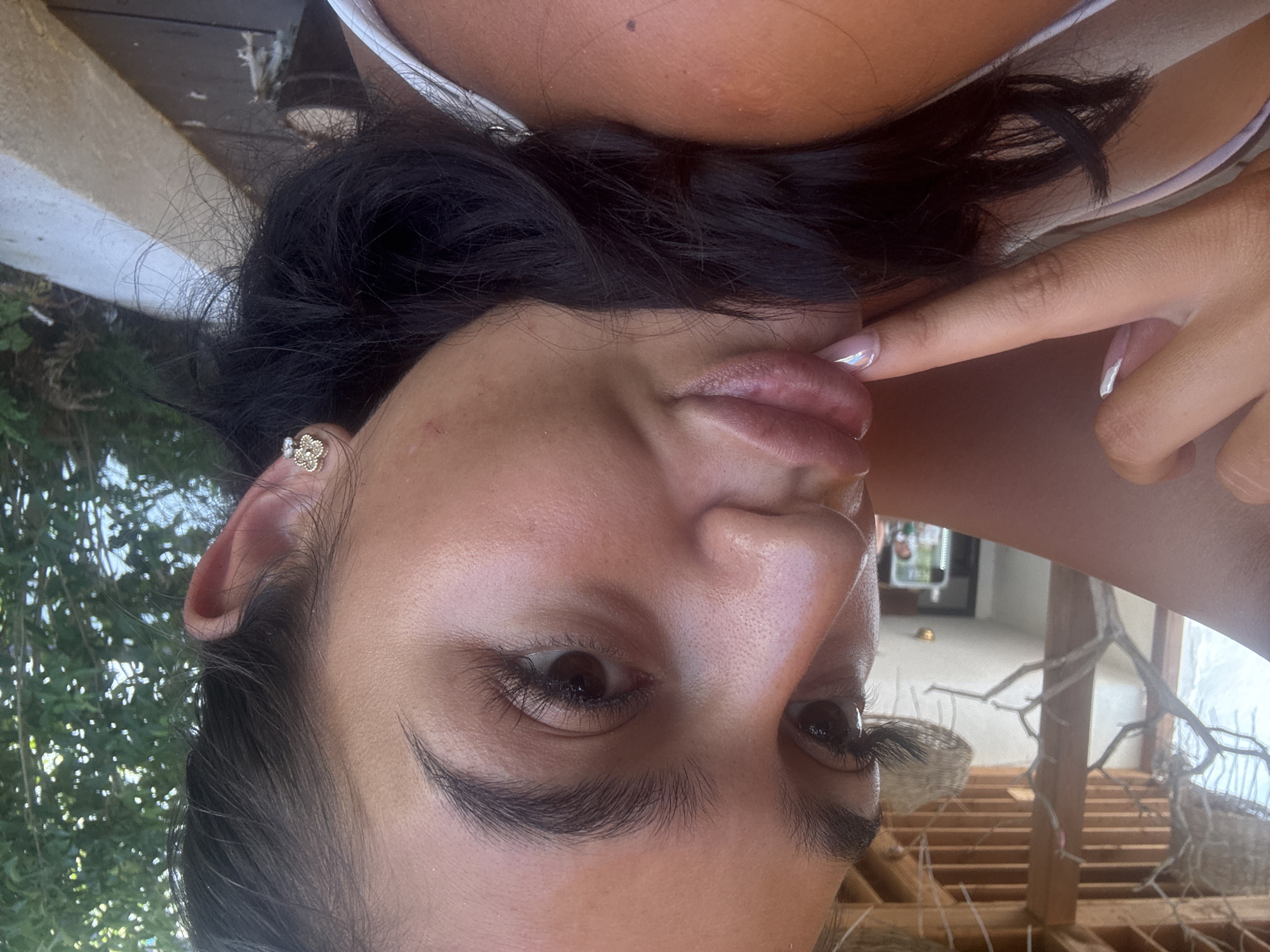 A close-up, over-the-shoulder selfie of a young woman with dark hair and thick eyelashes, wearing a white tank top and a small, jeweled earring. She is touching her lips with her finger, her nails painted with a french manicure. The background suggests an outdoor setting with wooden structures and greenery, possibly in Primorsko, Bulgaria.