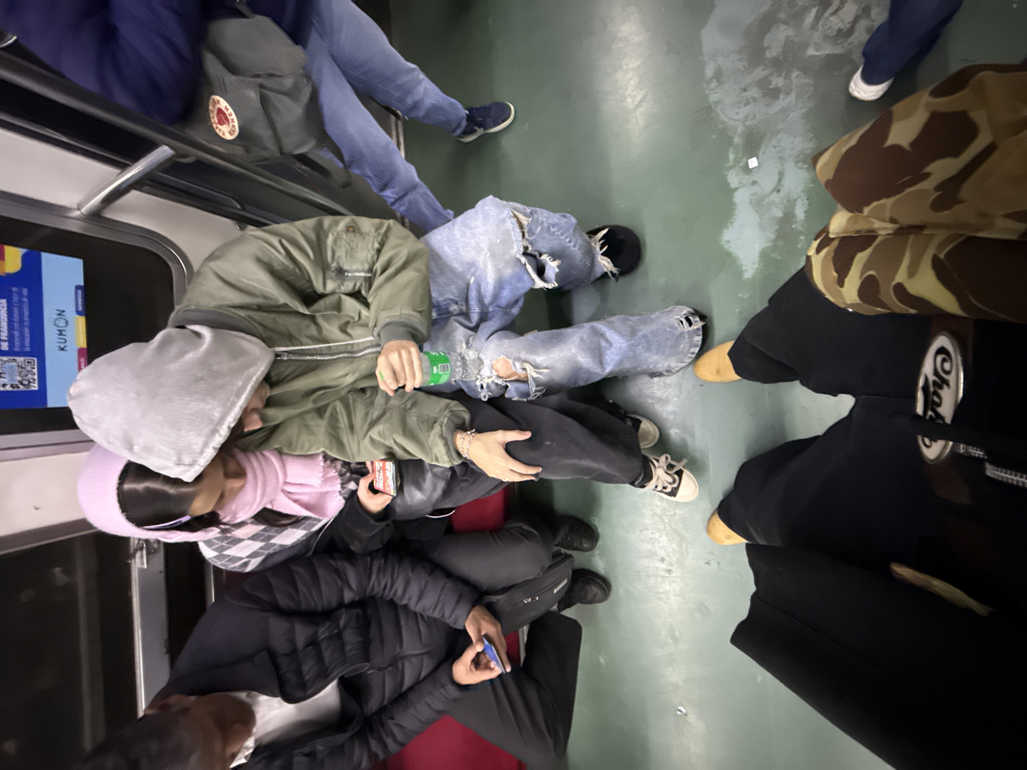 The image depicts the interior of a subway car, likely in Buenos Aires, Argentina, given the presence of the Spanish language and branding of 'KUMON' on the advertisement within the train car. Several individuals are present. The subjects are oriented in various directions. One person in blue jeans is seen sitting on the floor of the subway car, occupying considerable space. Another individual is dressed in a light green jacket, ripped blue jeans and black shoes. A third person, possibly a woman, wears a light pink hooded sweatshirt and is looking down at her phone. Another person has a black puffer coat. One person wears a camouflage jacket and black pants. Some passengers are seated while others stand, indicating different levels of comfort or space availability within the subway car. The environment appears to be a typical subway car interior with metal handrails, seating, and advertisements.
