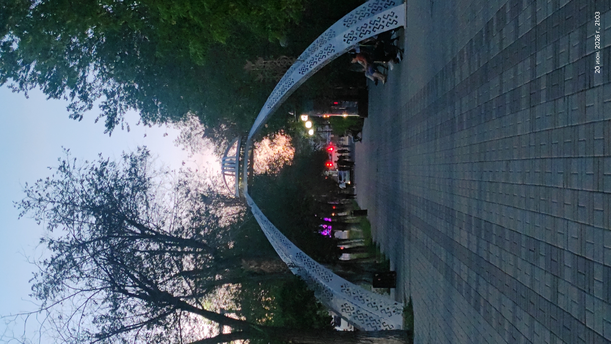 The image depicts a park-like setting in Astana, Kazakhstan, likely during twilight. A prominent arched structure with a decorative, possibly metallic, design spans across a paved walkway. Trees line both sides of the walkway, casting shadows and creating a canopy effect. The sky above is a pale blue with hints of pink and orange from the setting sun, visible through the arch and between the trees.
Several individuals are present in the scene. On the right side of the walkway, two people are seated on a bench, appearing relaxed. Further down the path, a group of people are walking, some silhouetted against the fading light. A few vehicles are parked or moving in the background, and streetlights are illuminated, casting a warm glow. The walkway itself is constructed of grey paving stones arranged in a pattern. The overall atmosphere is serene and peaceful, suggesting a public space used for recreation and transit.