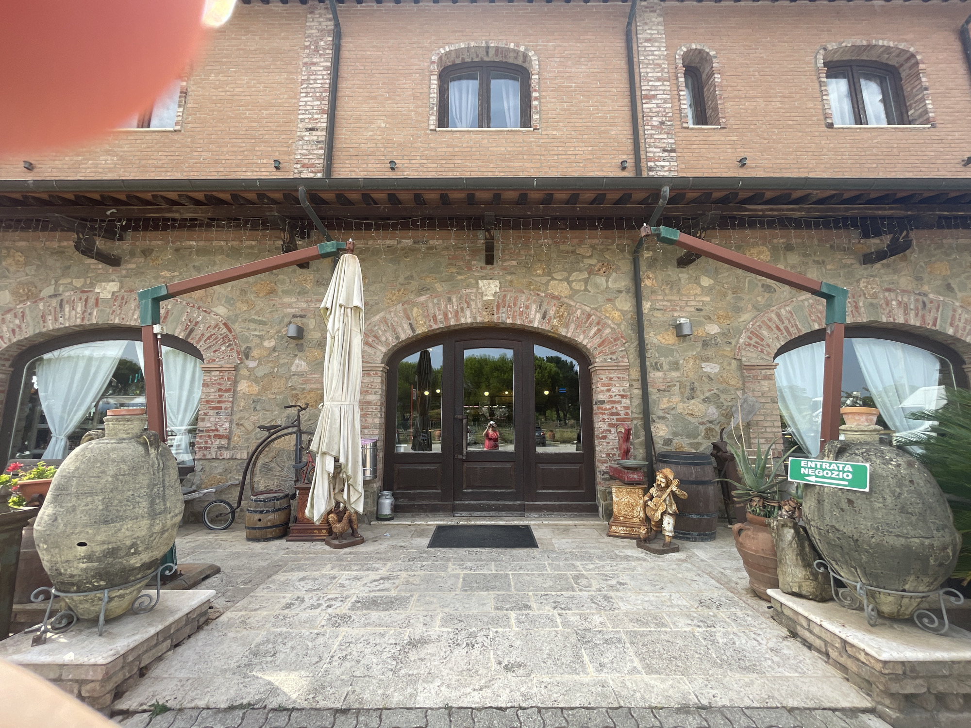 The image shows the entrance to a store in Follonica, Italy. The building has a brick facade with arched windows and doors. In front of the entrance, there are several large terracotta pots, a vintage bicycle, and two barrels. A sign that reads "ENTRATA NEGOZIO" (Shop Entrance) is visible on the right. The ground is paved with stone. A person in a red dress is reflected in the glass doors. The lighting suggests it is daytime, possibly late afternoon, with some sunlight present.