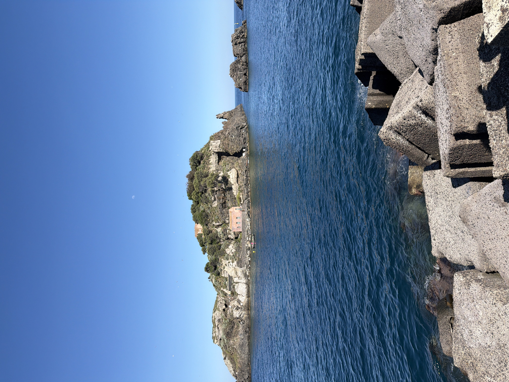 The image shows a view of Aci Castello, Italy. The scene includes a rocky coastline, a small island with buildings and vegetation, the blue sea, and a clear blue sky with a sliver of the moon visible. Large concrete tetrapods are situated on the right side of the frame, meeting the sea.