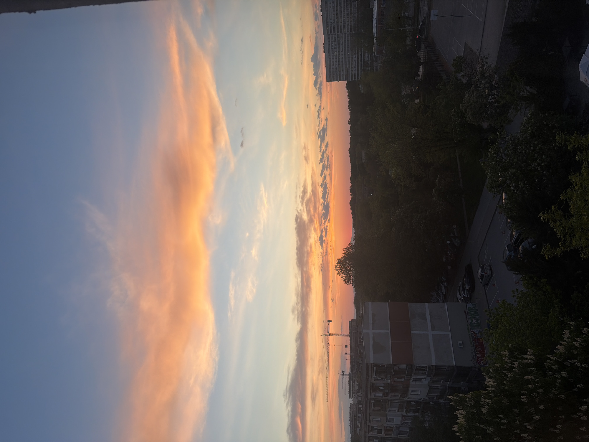 The image depicts a sunset view over a residential area in Plovdiv, Bulgaria. The sky is filled with colorful clouds in shades of pink, orange, and blue. Buildings and trees are visible in the foreground. A construction crane is also visible. Cars are parked on the side of a street. There are no people visible in the image.