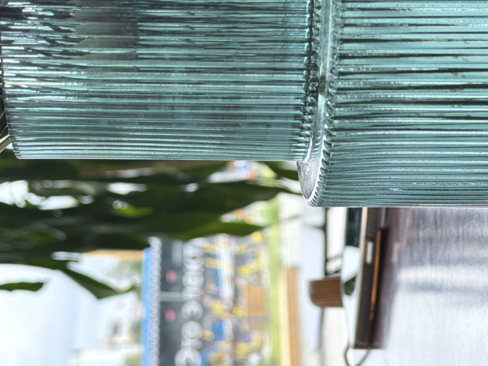 The image shows a close-up of a glass lampshade with horizontal ribbed detailing. The lampshade has a greenish-blue tint and is composed of stacked, thin glass panels. The background, seen through the glass, is blurred, but foliage suggests a garden or outdoor setting. Also visible in the background is a blurry display of items with letters and numbers, along with a building. There's also part of a wooden shelf, and a reflective surface. The exact location in Moscow, Russia, cannot be determined from this limited information.