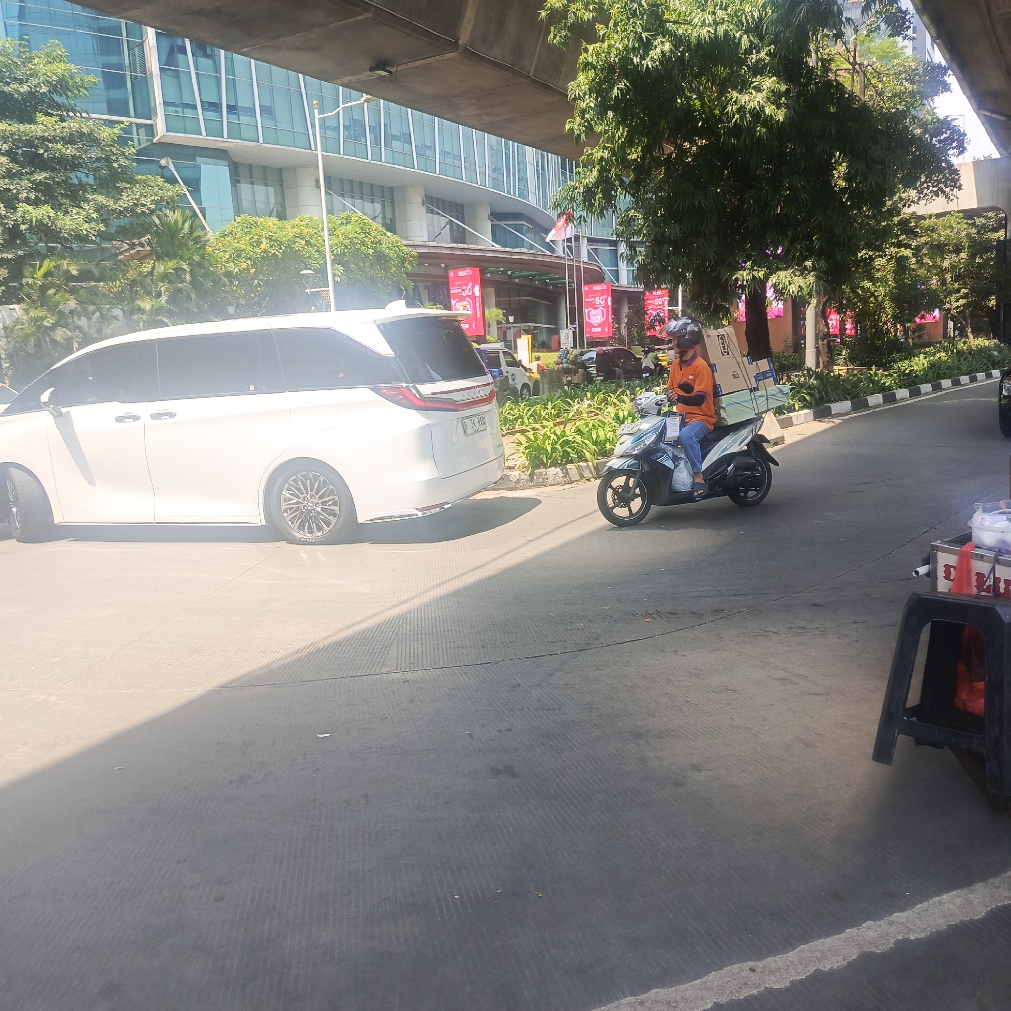 A delivery driver on a scooter with boxes strapped to the back is seen in front of a modern glass building. The driver is wearing an orange shirt and a helmet. A white minivan is parked to the left of the scooter. The scene appears to be on a street or parking lot in South Jakarta, Indonesia, as suggested by the architectural style and tropical trees. The ground is asphalt, and there is a white line marking a border. Sunlight casts shadows on the ground.