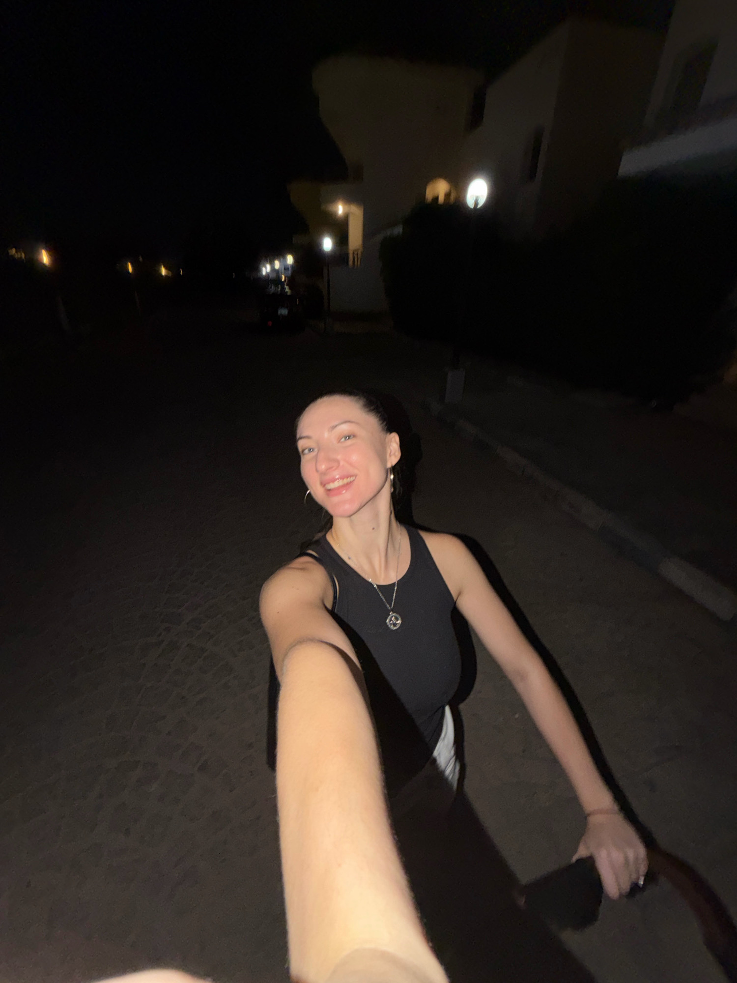 A young woman with dark hair pulled back is walking at night on a paved path or road in Sharm El Sheikh, Egypt. She is smiling and holding a camera or phone out in front of her, taking a selfie. She is wearing a black tank top and white shorts, and a necklace with a circular pendant. The background is dark, with some building lights visible in the distance, suggesting a residential or commercial area. The lighting is dim, creating shadows and a nighttime atmosphere.