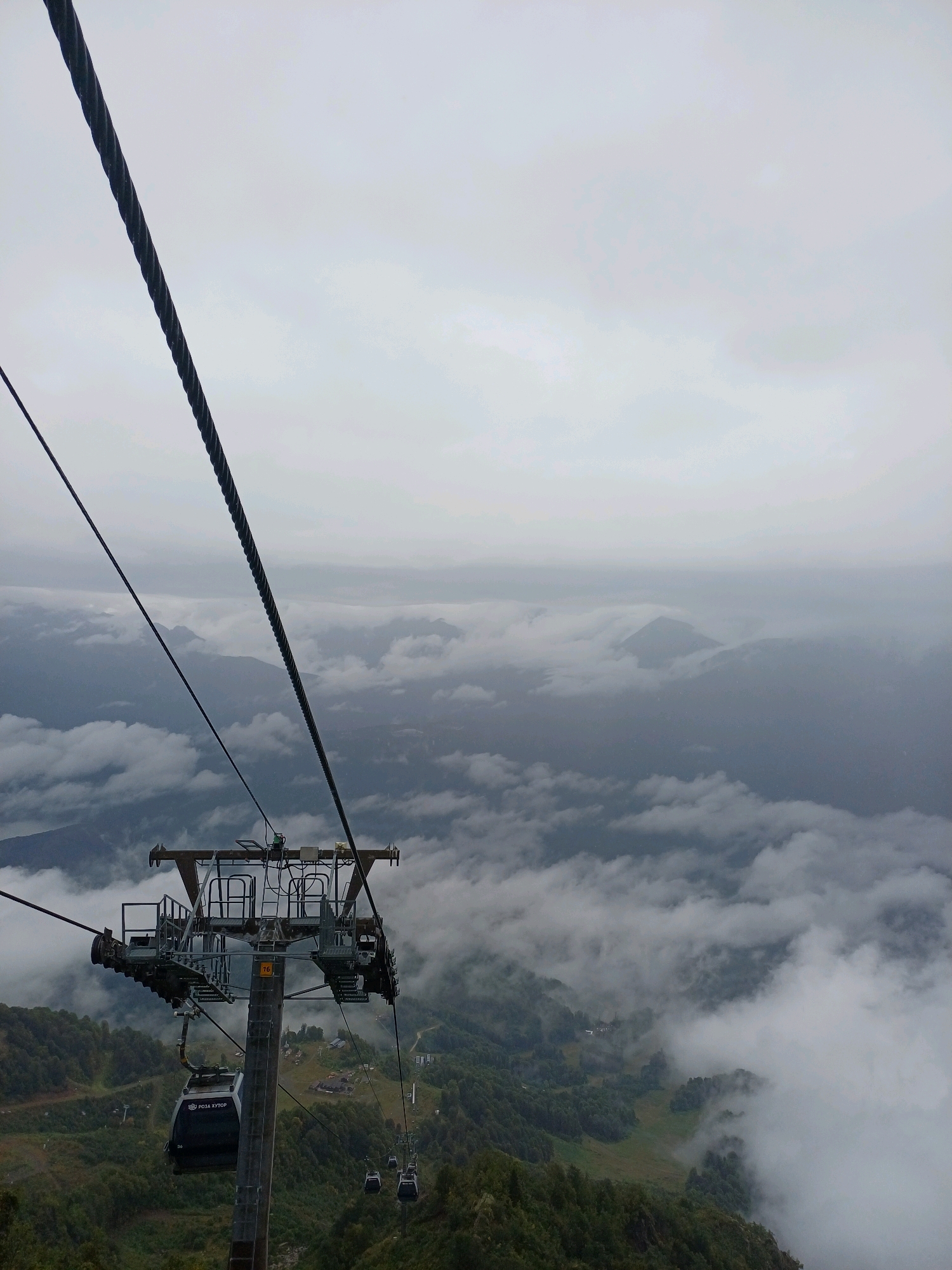 The image shows a cable car in motion, high above a mountainous landscape shrouded in clouds. The cable car is suspended from thick steel cables that stretch across the frame, disappearing into the hazy sky. In the foreground, the intricate metal structure of the cable car station is visible, with a dark, modern cable car attached and descending. Below, the lush green slopes of the mountains are dotted with trees and small buildings, suggesting a settlement or resort area. The presence of other cable cars further down the line indicates an active transportation system. The overall atmosphere is one of elevation and a vast, misty expanse, hinting at the location in Estosadok, Russia, known for its ski resorts and mountain views. The weather appears overcast, with a soft, diffused light.