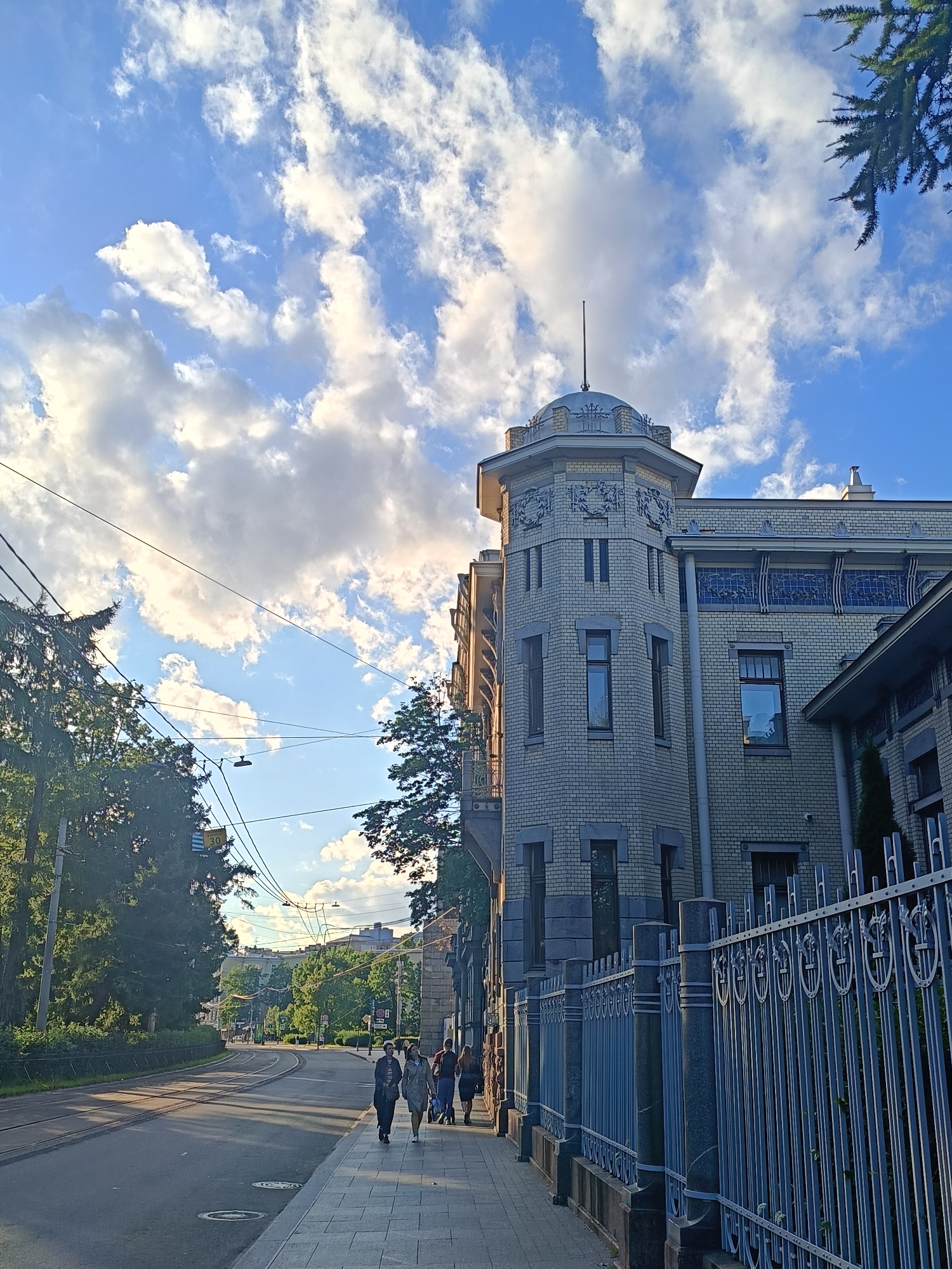 The image depicts a street scene in Saint Petersburg, Russia, with a prominent Art Nouveau building on the right side. The building is constructed of light-colored brick with ornate decorative elements, including a domed turret and stained-glass windows. A metal fence with intricate patterns runs along the sidewalk in front of the building. On the left side of the street, trees and overhead wires are visible. A tram track runs down the center of the asphalt road. Several people are walking on the sidewalk, appearing to be engaged in casual movement. The sky is blue with scattered white clouds, and the lighting suggests it is either morning or late afternoon. The overall atmosphere is urban and somewhat historic.