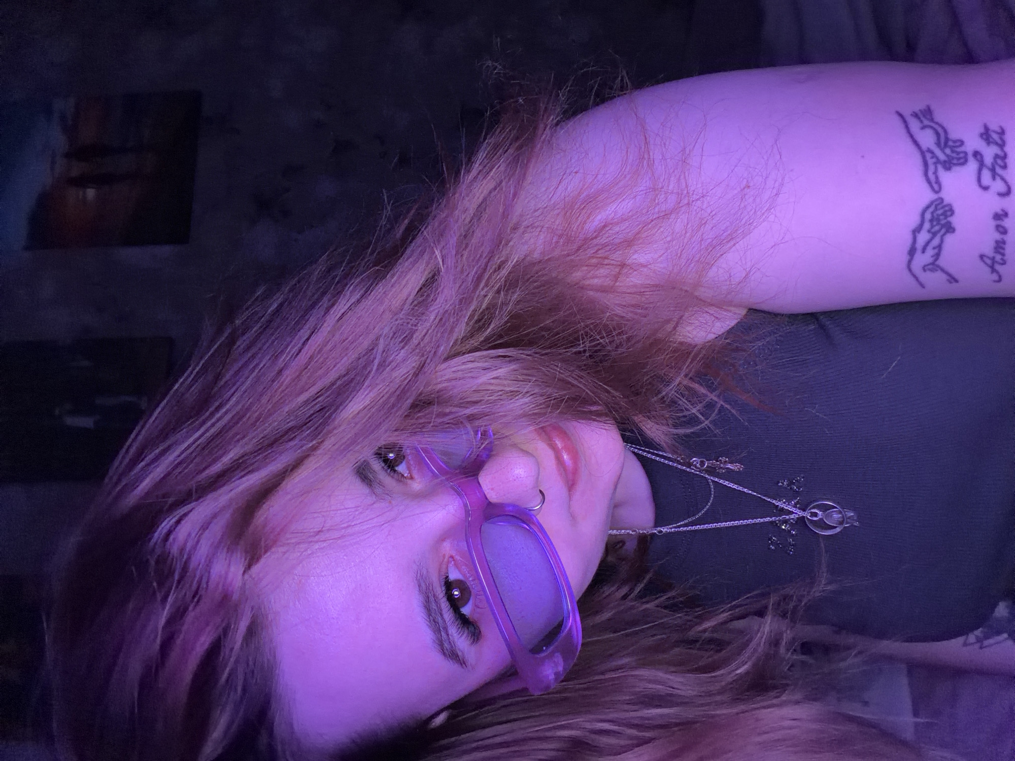 A young adult woman lies on her back, with her head tilted to the left. She has reddish-brown hair and is wearing lavender-tinted sunglasses. She also wears a dark green top and two silver necklaces. She has a tattoo of two hands reaching out to each other with the text "Amor Fati" on her right forearm. The background is dark, with what appears to be a picture or poster on the wall. No location can be determined.