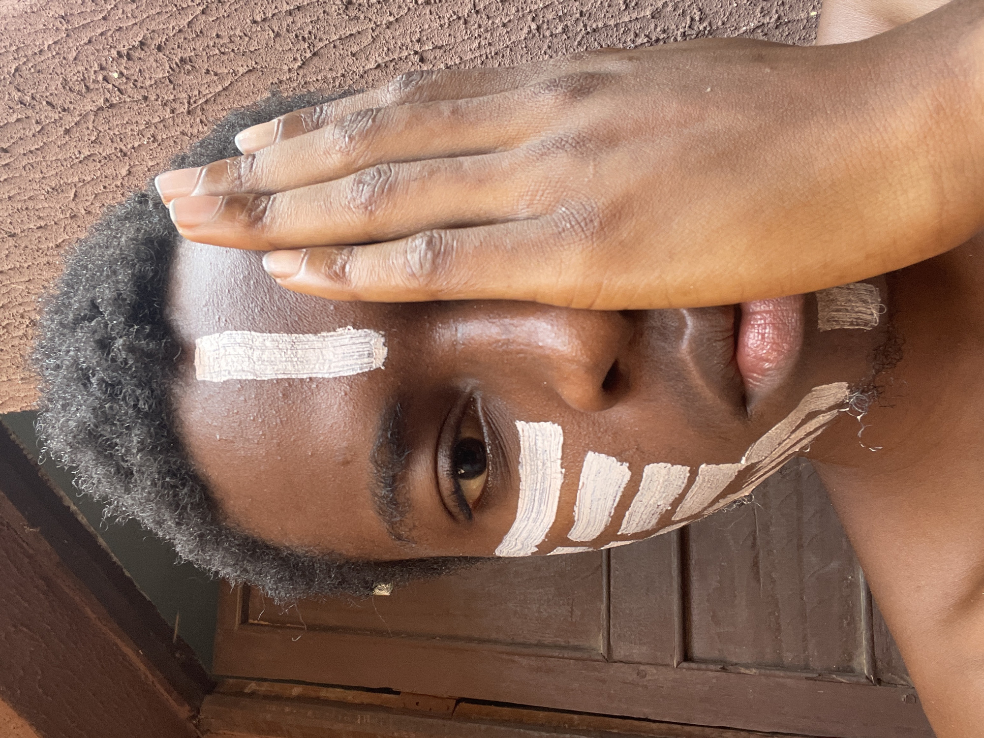 This image, taken in Onitsha, Nigeria, captures a close-up portrait of a person with dark skin, oriented vertically (rotated 90 degrees counter-clockwise from a standard portrait). The subject's right hand is raised, covering a significant portion of their upper face, including the forehead and nose. White, linear paint markings are visible on the exposed areas of the face: a thick horizontal stripe across the left forehead and several vertical stripes on the right cheek, below the eye.
The left eye is open and appears to look directly at the viewer with a neutral or contemplative expression. The person has short, dark, curly hair and some sparse facial hair. The background features a textured, reddish-brown surface, possibly a wall, in the upper part of the frame, and a darker brown, segmented wooden structure in the lower part. The lighting is bright and natural, suggesting it is daytime. The presence of face paint indicates a potential cultural practice, artistic expression, or celebratory event. No specific activity other than posing is discernible, and there is no visible text in the image.