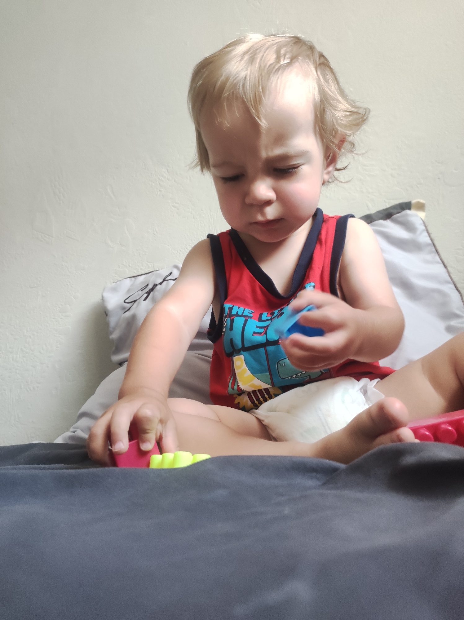 A fair-haired toddler, wearing a red tank top with blue trim and a dinosaur graphic, is sitting on a gray blanket with pillows behind them. The toddler is looking down and playing with colorful building blocks, holding a blue one in their right hand and interacting with a pink and yellow block with their left. The toddler is also wearing a diaper. The background consists of a textured off-white wall. There is no visible indication of the city of Chaikovsky, Russia, in the image.