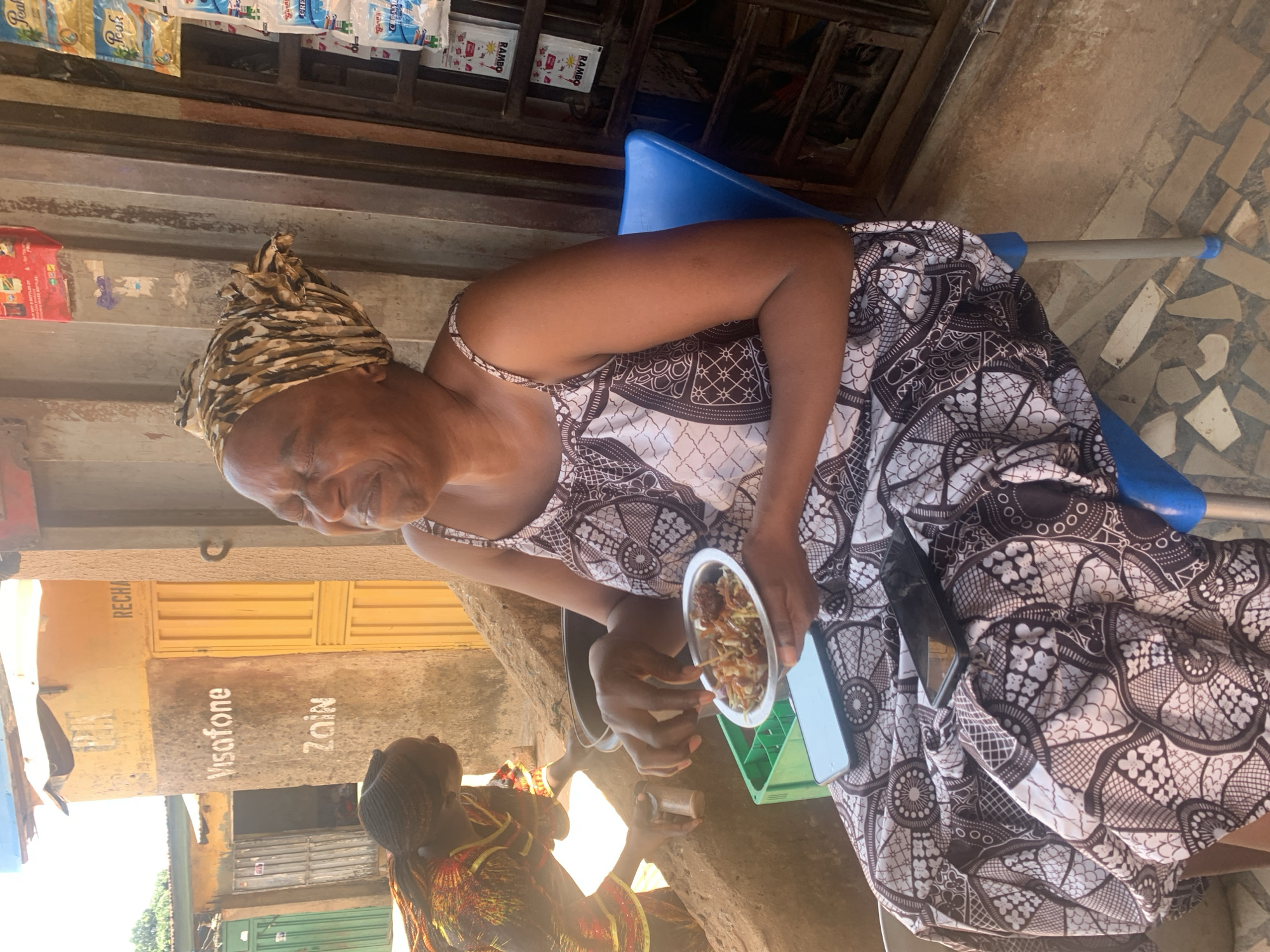 An elderly woman with a headscarf is seated on a blue plastic chair in Sabon Tasha, Nigeria, eating from a metal bowl. She is wearing a patterned dress. A smartphone and a green plastic holder are in her lap. Another person is visible in the lower-left corner of the image. The background includes a storefront with advertisements for "Visafone" and "Zain".
