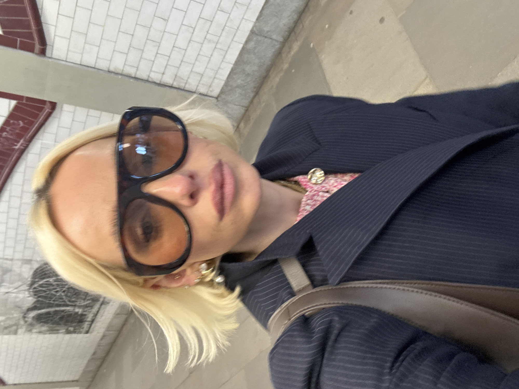The image displays a close-up, selfie-style photograph of an adult individual with short, light blonde hair. The subject is wearing large, dark tortoiseshell-patterned sunglasses with amber-tinted lenses, a dark blue pinstriped blazer, and a pink and white textured top underneath, adorned with a circular gold-colored pendant. Multiple earrings, including a prominent pearl-like drop earring, are visible on the subject's left ear. A brown leather shoulder strap is seen over the subject's left shoulder.
The background indicates an indoor, subterranean environment consistent with a pedestrian tunnel or platform of the London Underground in London, United Kingdom. The walls are constructed from white rectangular tiles, featuring a distinctive horizontal dark red tiled trim. On the left side of the frame, a large, dark, abstract artwork, appearing to be composed of interwoven metallic structures, is partially visible against the tiled wall. The floor consists of large, light grey stone or concrete slabs. The subject's lips are slightly parted, and their expression is neutral, largely obscured by the sunglasses. No other individuals are present in the frame.