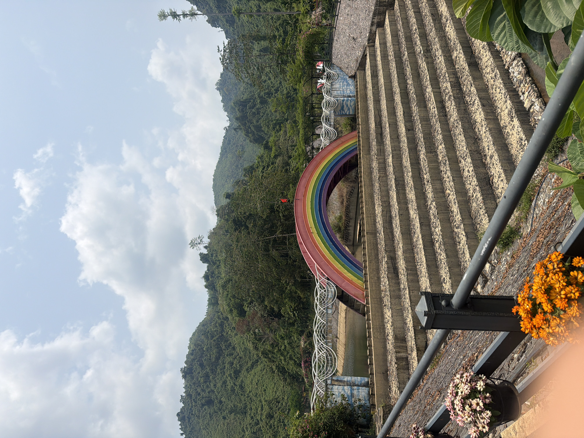 The image depicts a vibrant rainbow-colored structure set against a backdrop of lush green mountains and a partly cloudy sky. The rainbow forms an arch over what appears to be a waterway or reservoir, supported by white decorative elements. To the right, a tiered concrete structure adds an element of architectural design. A metal railing with hanging flower pots is visible in the foreground. The location is most likely Ba Na Hills near Đà Nẵng, Vietnam.