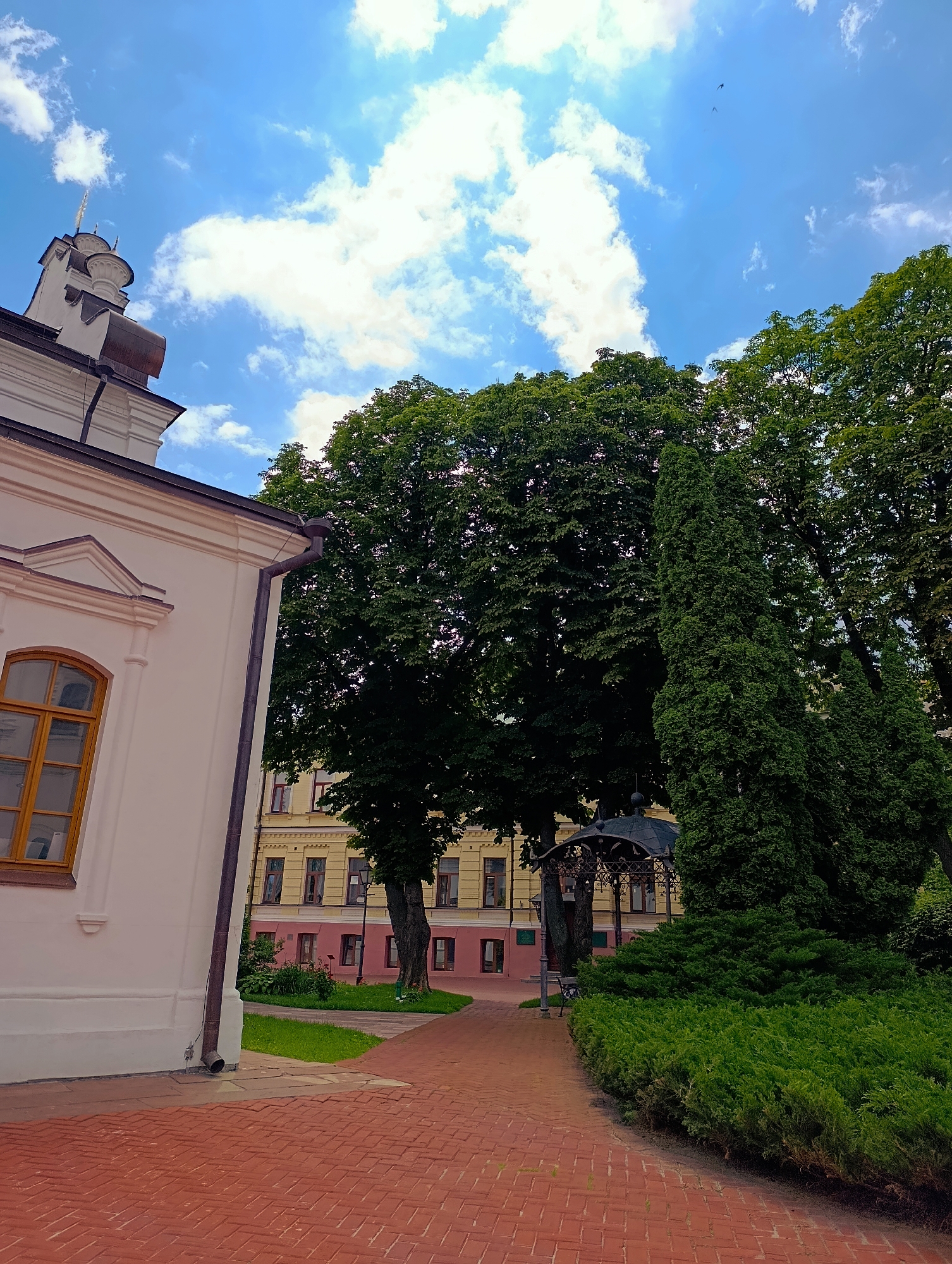 This is a low-angle, eye-level, outdoor, daytime, full shot of a park area in Kyiv, Ukraine, near a building with white walls and a prominent arched window with a wooden frame. The building's roofline and a dome-like structure are visible at the top left. The ground is paved with red bricks in a herringbone pattern. A red brick pathway curves to the right, bordered by dense green bushes and tall, conical evergreen trees. Beyond the bushes, a dark gazebo with an ornate metal roof is visible, and further back, a yellow building with a pink base and multiple windows stands. Large, leafy trees with dark green foliage dominate the background, obscuring much of the sky, which is a bright blue with scattered white clouds. Two small, dark shapes, possibly birds, are visible in the sky near the top center of the image. A dark drainpipe runs down the side of the white building, with a downspout at its base.