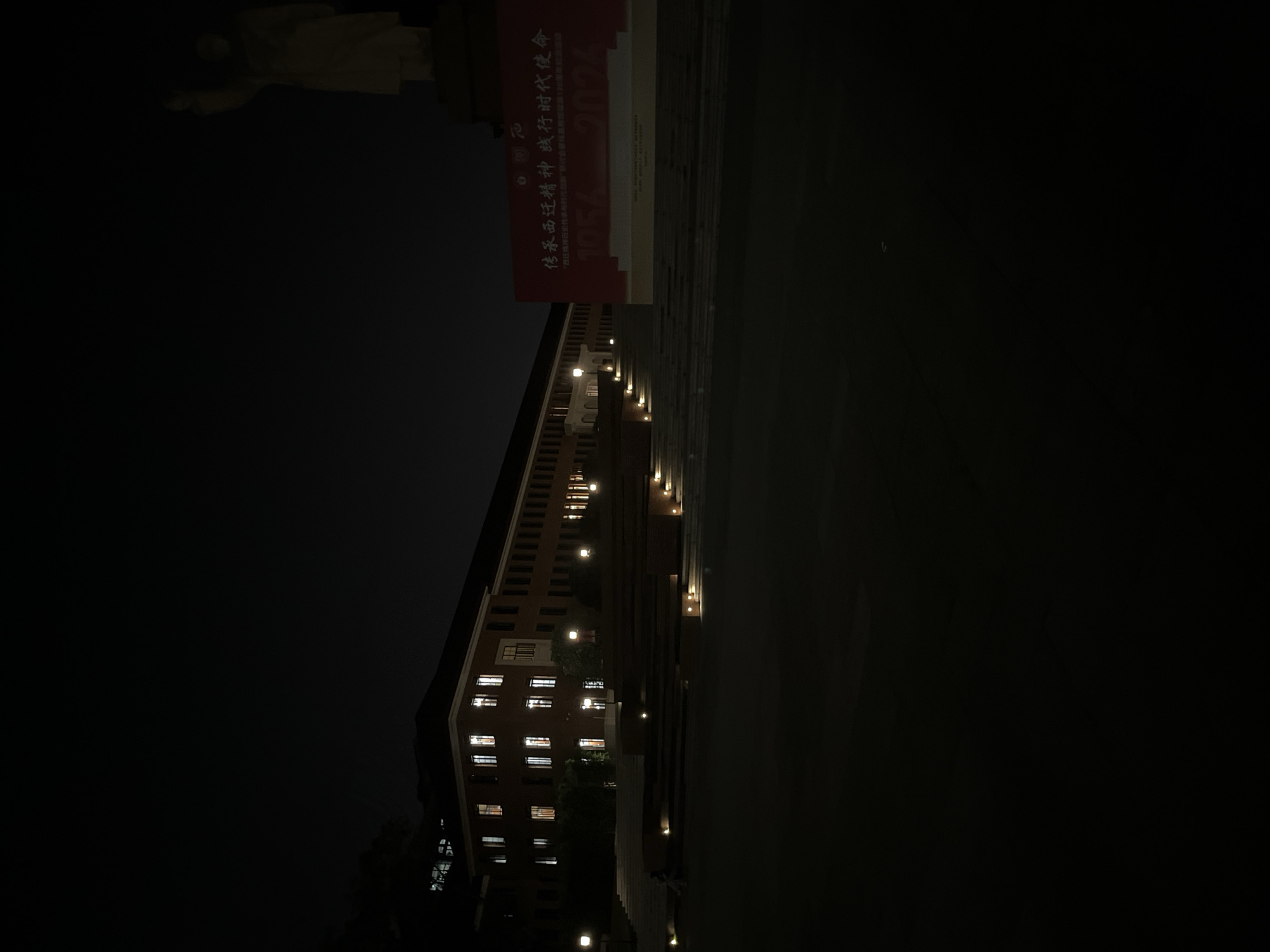 The image captures a tall building in Yuzhong District, China, illuminated at night. The building features a repetitive window pattern with interior lights visible, and small exterior lights run vertically along its facade. A statue stands in the background, above a red sign with Chinese characters. The sign reads "传承西迁精神 践行时代使命 '西迁精神历史传与时代创新 研讨会旗120周年纪念活动' 1056 2026". The surrounding environment appears dark.