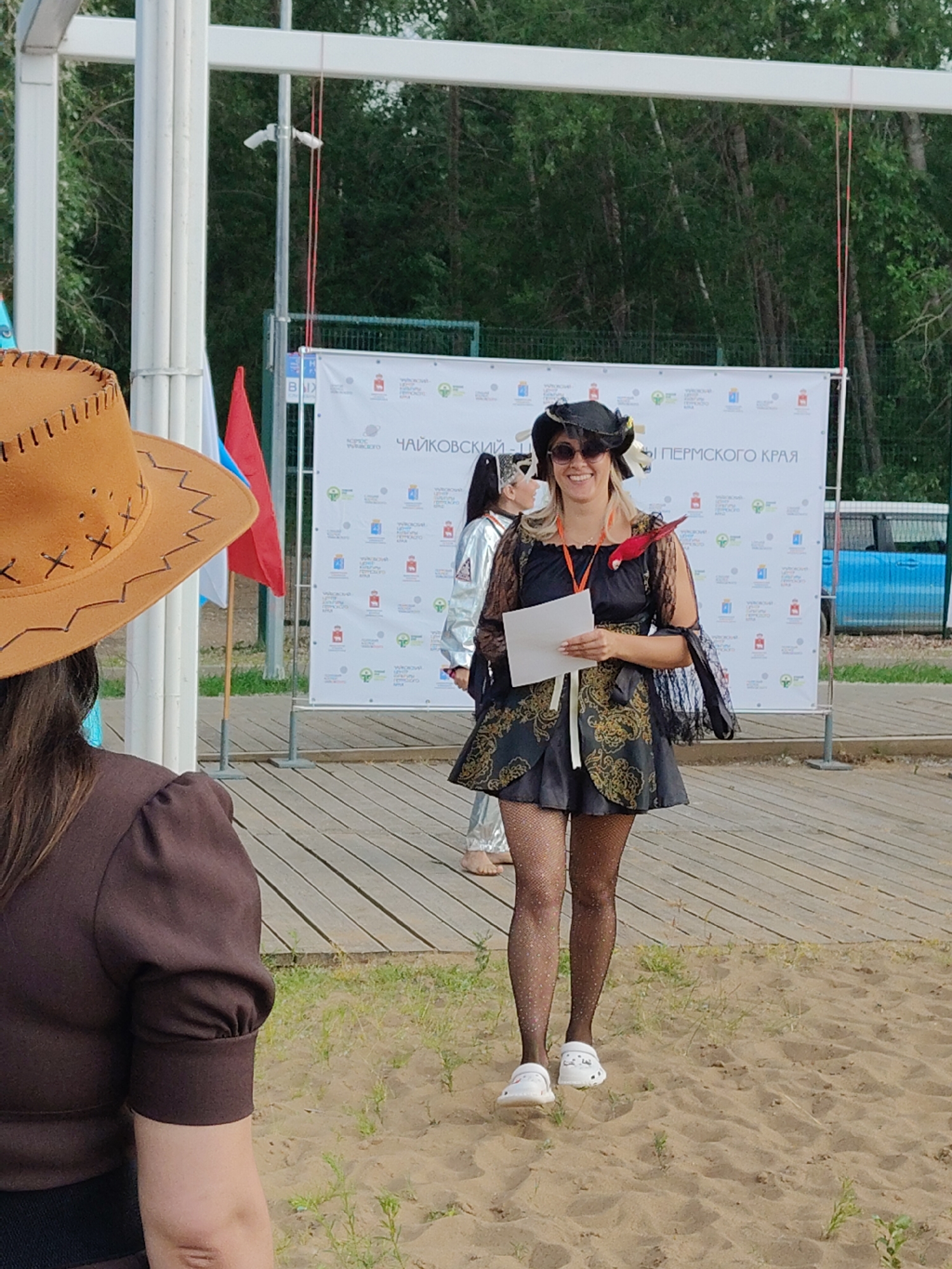 In an outdoor setting, a woman wearing a black pirate-themed costume with a ruffled dress, fishnet stockings, and a black hat stands on a sandy area. She holds a piece of paper and smiles at the camera, with a red parrot perched on her shoulder. She is wearing white Crocs. To her left, another woman in a silver outfit stands partially visible, and a person wearing a brown shirt and a cowboy hat is in the foreground, with their back to the camera. Behind them, a large banner with text and logos is displayed. A wooden walkway runs behind the sandy area, and trees are visible in the background. A blue vehicle is parked to the right of the banner.
