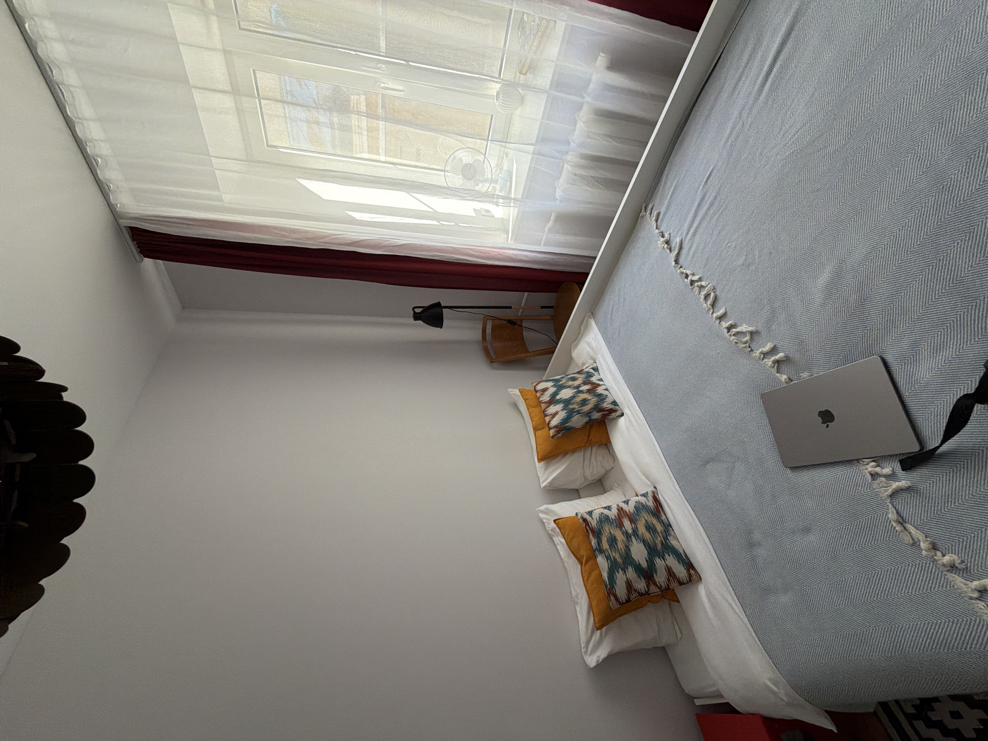 A medium shot captures a bedroom scene, likely in an apartment in Saint Petersburg, Russia, featuring a neatly made bed covered with a light blue blanket and white sheets. Two decorative pillows with geometric patterns rest at the head of the bed, complementing two plain orange pillows. A laptop computer is positioned open on the bed, suggesting someone was recently working.
The room is illuminated by natural light streaming through a large window draped with sheer white curtains and accented by dark red drapes. The window reveals a glimpse of buildings and a radiator. To the left of the window, a black floor lamp stands next to a wooden chair. The walls are painted a neutral, light gray, and a dark, decorative light fixture hangs from the ceiling in the upper left corner of the frame. A sliver of a red bedside table is visible on the far left. The flooring is partially visible in the lower left corner, showing a black and white patterned rug. The overall impression is one of a well-lit, possibly cozy, personal space.