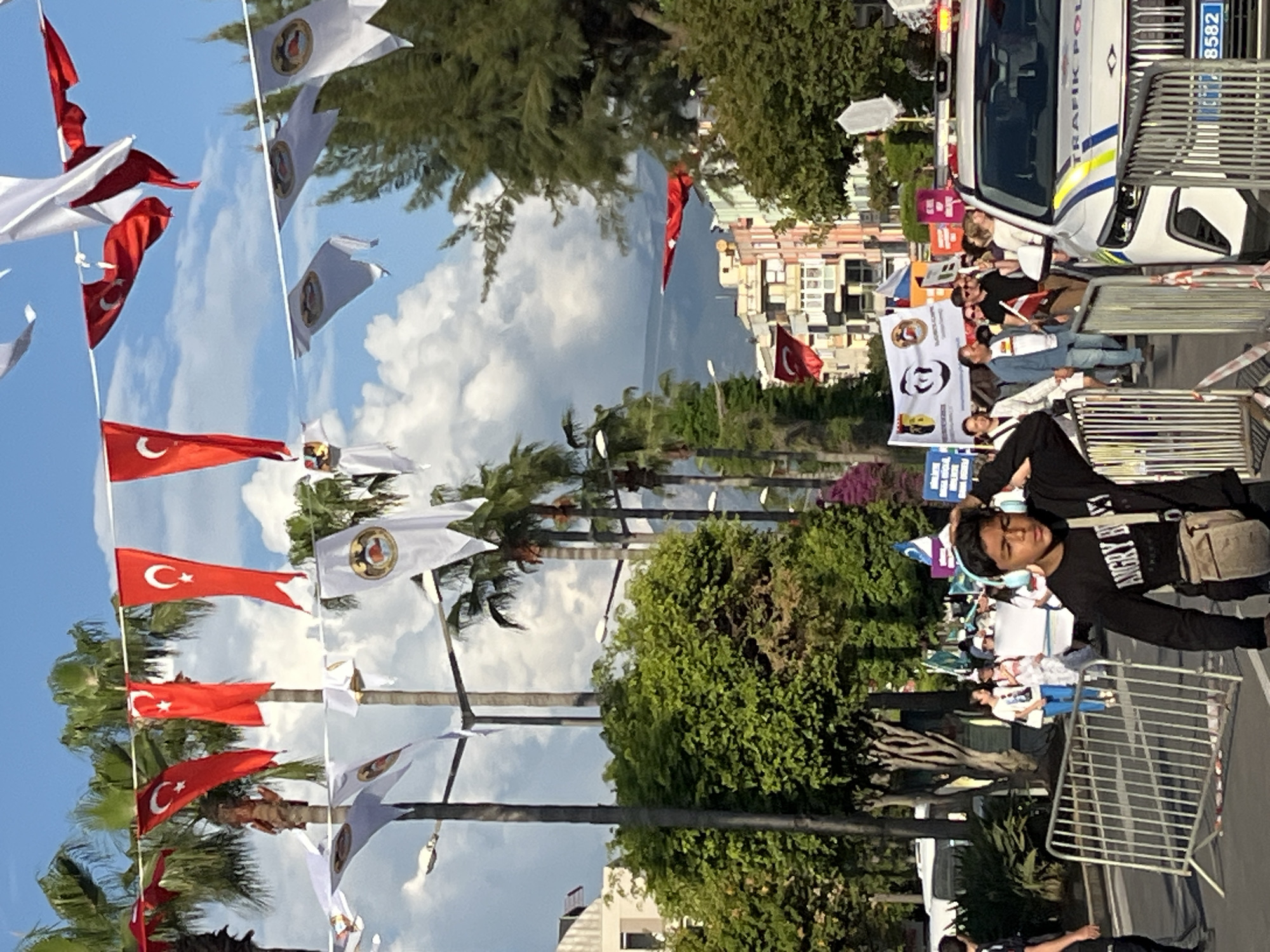 Here is a description of the image:
The image shows a scene from what appears to be a street in Alanya, Turkey. In the foreground, a young person wearing headphones is visible, with a bag slung over their shoulder. Metal barricades are present, suggesting a controlled environment. The background reveals a gathering of people, some holding banners, possibly indicating a demonstration or event. Turkish flags are prominently displayed along the street, hanging from poles lined with palm trees. A police vehicle is visible, its lights not active, hinting at a presence for security purposes. Buildings are visible in the background, defining the urban landscape of Alanya. The sky is clear with some clouds visible.