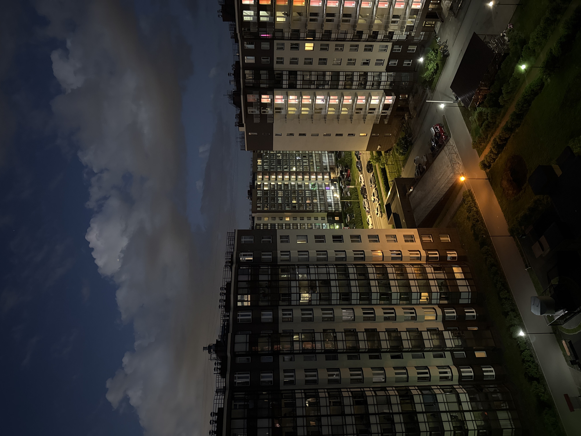 An aerial night view of a residential complex in Saint Petersburg, Russia, depicts several multi-story buildings with illuminated windows, suggesting occupancy. The architecture features modern designs with balconies and dark-colored facades contrasted by light-colored sections. Cars are parked along a road next to one of the buildings, and streetlights cast a warm glow on pathways and green spaces. The sky above is dark blue with scattered clouds, and a faint crescent moon is visible. The scene conveys a sense of urban tranquility under the night sky.