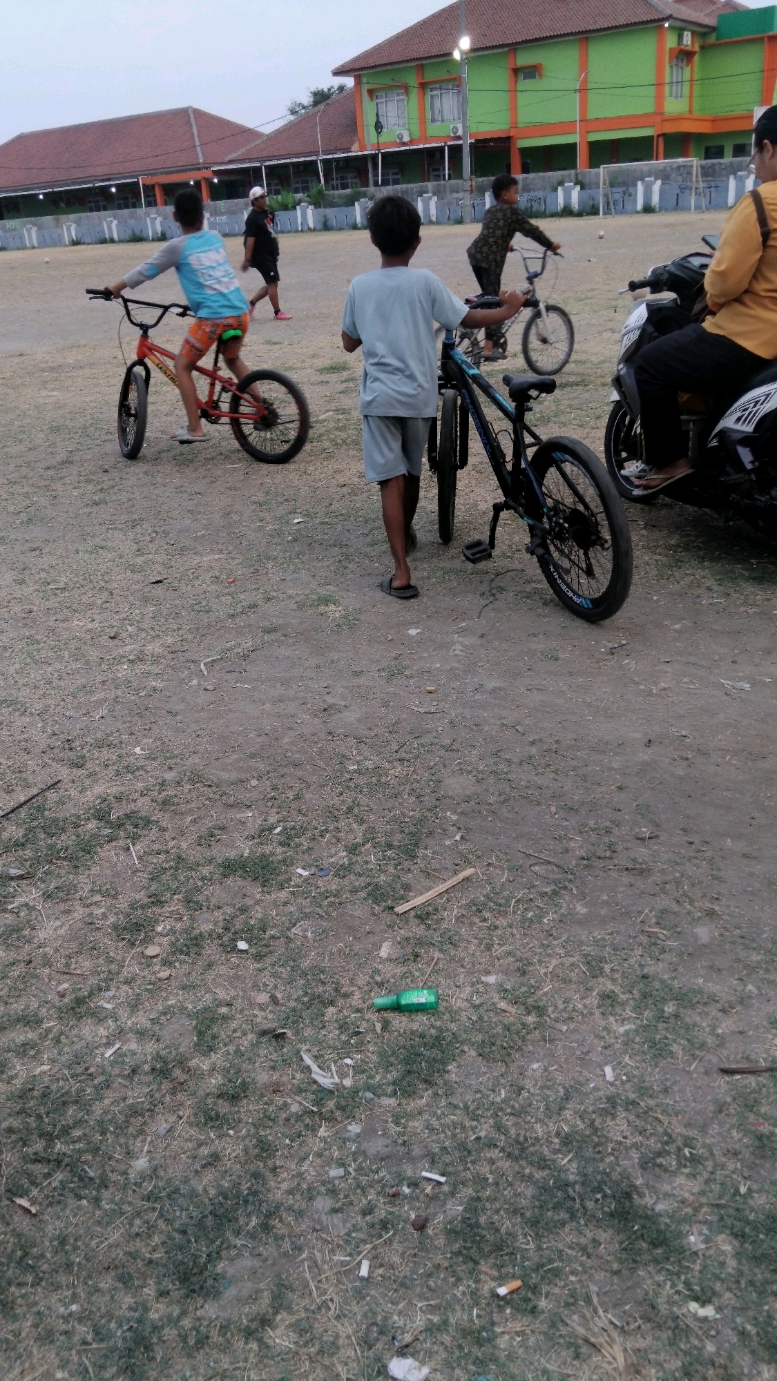 The image depicts an outdoor scene in Tegal, Indonesia, possibly a schoolyard or a recreational area, judging by the presence of buildings in the background that resemble educational or sporting facilities. The ground is covered with dry grass and dirt, with scattered debris including cigarette butts and a green plastic bottle.
In the foreground, a young boy is walking next to a black bicycle. The bicycle is a mountain bike with a blue rim on the rear wheel and a black rim on the front wheel. The boy is wearing a light blue t-shirt and grey shorts.
Behind him, another boy is riding a red BMX-style bicycle. He is wearing a blue long-sleeved shirt with a white graphic design and orange shorts. His bicycle has red rims on both wheels and appears to be a smaller frame.
Further in the background, several other individuals are present. One person in a dark outfit is standing near a white bicycle. Another person, wearing a patterned outfit, is also on a bicycle. To the right of the scene, a black motorcycle is parked, and a person wearing a yellow shirt is seated on it. The buildings in the background are painted in green and orange, and there are structures resembling goalposts in the distance, suggesting a sports field. The sky is overcast.