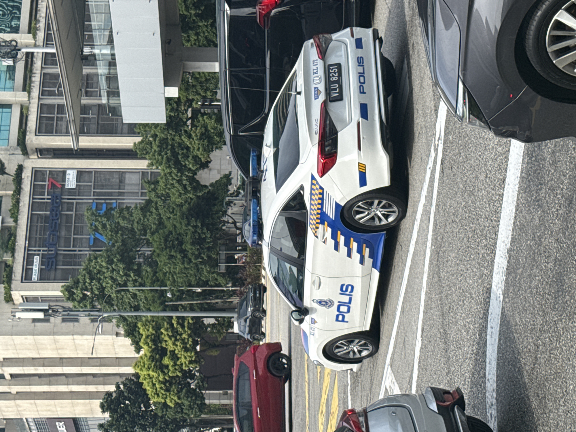 The image captures a vibrant urban street scene in Kuala Lumpur, Malaysia, dominated by a white Malaysian police car during bright daylight. The police vehicle, a Honda Civic, features distinctive blue and yellow checkered patterns and stripes along its side and rear. The word "POLIS" is clearly visible in blue lettering on both the car's side and trunk. The license plate "VLU 8251" is displayed, with an additional plate below it showing "KL 472," indicating its registration in Kuala Lumpur.
Other vehicles are present, including a dark SUV parked behind the police car, a dark grey car to its immediate right, and a red sedan further down the street on the left. In the background, modern multi-story buildings with numerous windows line the street, partially obscured by green trees. Visible text on the buildings includes "JER," "PEJABAT URUSAN" (Management Office), "882654-D," and partially visible words like "TY" and "SOSONS". The road surface is asphalt with white and yellow markings. The clear lighting suggests it is daytime with good weather.