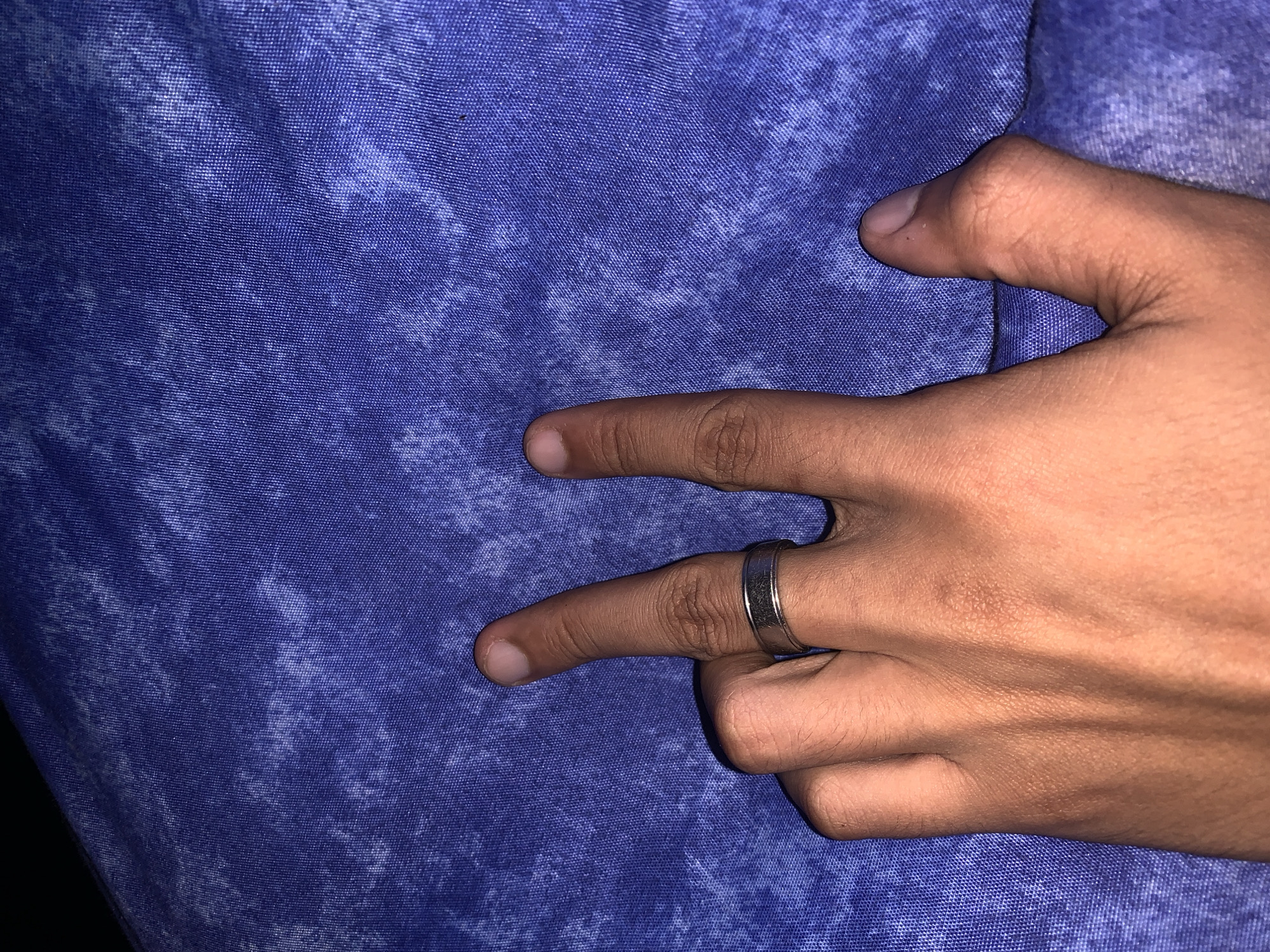 A close-up shot features a human hand against a textured blue fabric background, likely taken in Santa Ana, Venezuela. The hand, with a light to medium brown skin tone, is making a "V" sign or peace gesture, with the index and middle fingers extended upwards and the other fingers curled. A simple, metallic silver ring is worn on the ring finger of the hand. The blue fabric has a distinct mottled pattern, resembling a tie-dye or acid-washed effect, with lighter blue and white areas interspersed over a darker blue base. The weave of the fabric is clearly visible, suggesting a durable material. There are no other visible subjects, objects, or discernible activities beyond the hand gesture. No text is visible in the image. The lighting is somewhat soft, but the specific time of day or weather conditions cannot be determined.