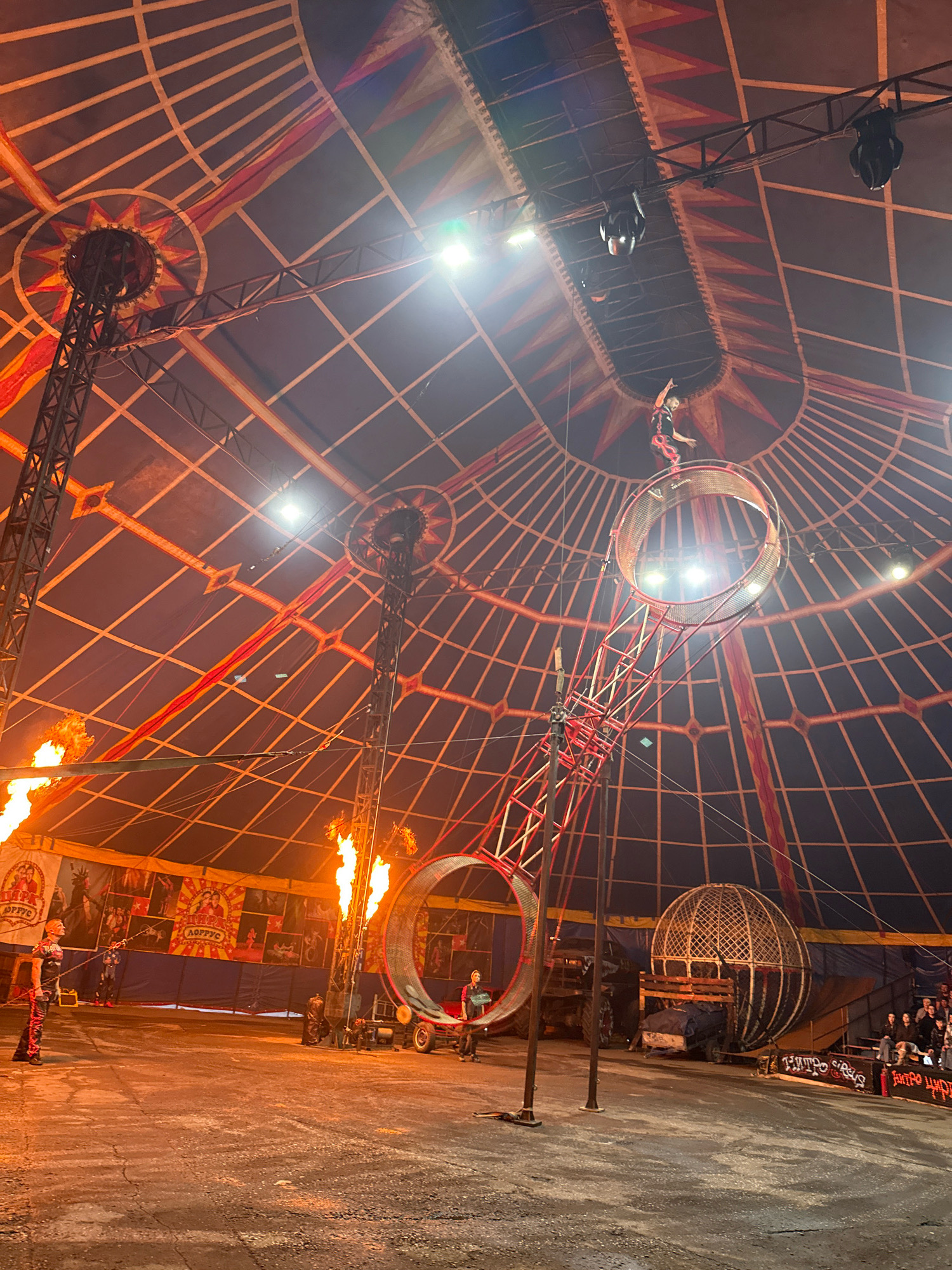 This image captures a scene inside a circus tent, likely located in Rusanovo, Russia, given the cyrillic text visible on banners. The tent has a domed ceiling with a grid-like structure of dark fabric crisscrossed by red and yellow decorative elements. Several spotlights are mounted on the ceiling structure.
In the center of the frame, a large, metallic, ring-shaped structure is elevated by a red metal framework. A performer, dressed in dark clothing with red accents, is positioned on top of this ring, appearing to be in mid-performance with one arm raised. Flames erupt from two large sources on the left side of the image, illuminating the lower section of the tent.
Further back, a large, spherical cage, resembling a "globe of death," is visible. Several individuals are seated in bleachers on the right side of the image, watching the performance. Various circus equipment and banners with cyrillic text, including images of performers, are present in the background. The ground appears to be a dark, dusty surface. The overall atmosphere suggests an active circus performance with elements of danger and spectacle.