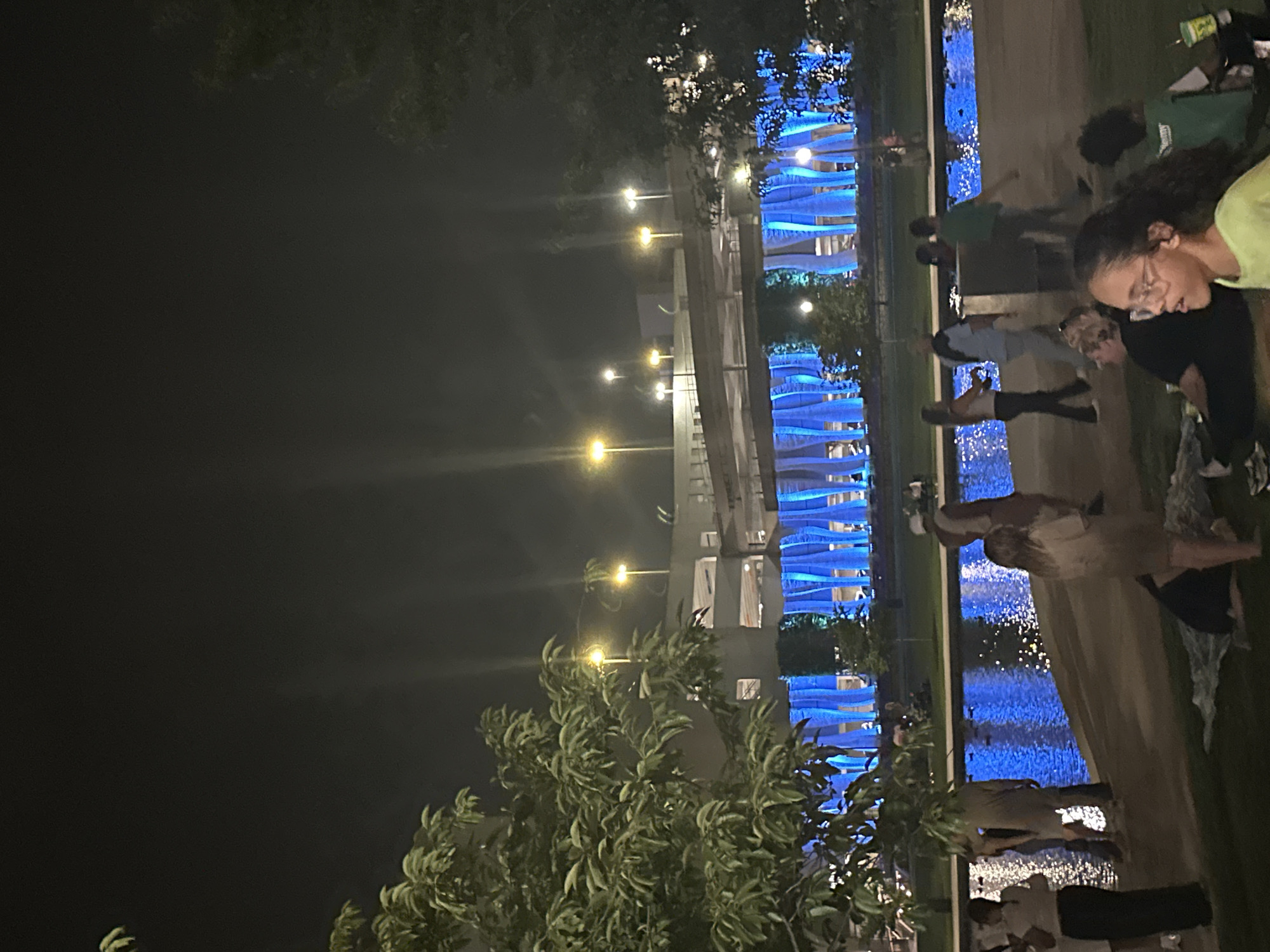 The image depicts a nighttime scene in downtown Huntsville, Alabama, likely at the Big Spring Park area. A crowd of people is gathered in front of a body of water, which reflects the vibrant blue lights illuminating the architecture behind it. The architecture appears to be a multi-story structure, possibly a parking garage or a building with a decorative facade, featuring vertical blue lights that create a striking visual effect.
Several individuals are visible in the foreground and mid-ground. On the left, a woman in dark clothing stands by the water's edge. Further back, a group of people are milling about, some sitting on the grass and others standing. A young girl with glasses is in the right foreground, looking to her left. The water itself is calm, with ripples and reflections of the blue lights and the overhead streetlights. Trees with green foliage are present on the left and right edges of the frame, framing the scene. The overall atmosphere is one of a public gathering or event at night.