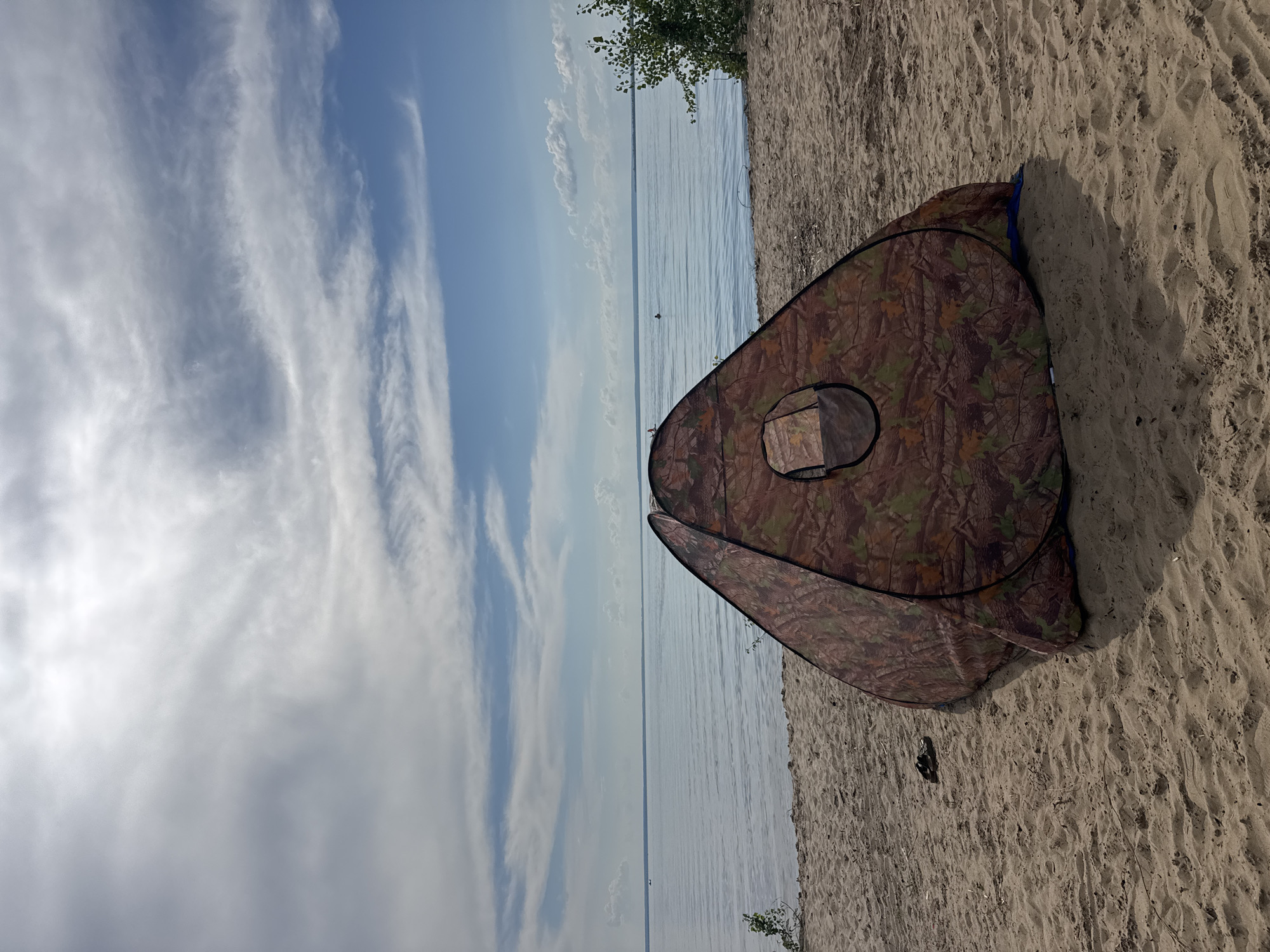 A camouflage pop-up tent is set up on a sandy beach. The tent is open, revealing a circular opening on one side. The tent is positioned on the right side of the frame, with its entrance facing away from the viewer. The beach sand is light brown and textured with footprints. Beyond the beach, a calm body of water stretches to the horizon, reflecting the sky. The sky is a vibrant blue with wispy white clouds that create a dynamic pattern. A few distant trees are visible on the far left of the frame, adding a touch of greenery to the scene. The lighting suggests it is daytime, likely late morning or early afternoon, with clear and bright conditions. There are no people visible in the frame. The scene evokes a sense of tranquility and relaxation, suggesting a peaceful day at the beach. The location is unclear but appears to be a natural, outdoor setting.