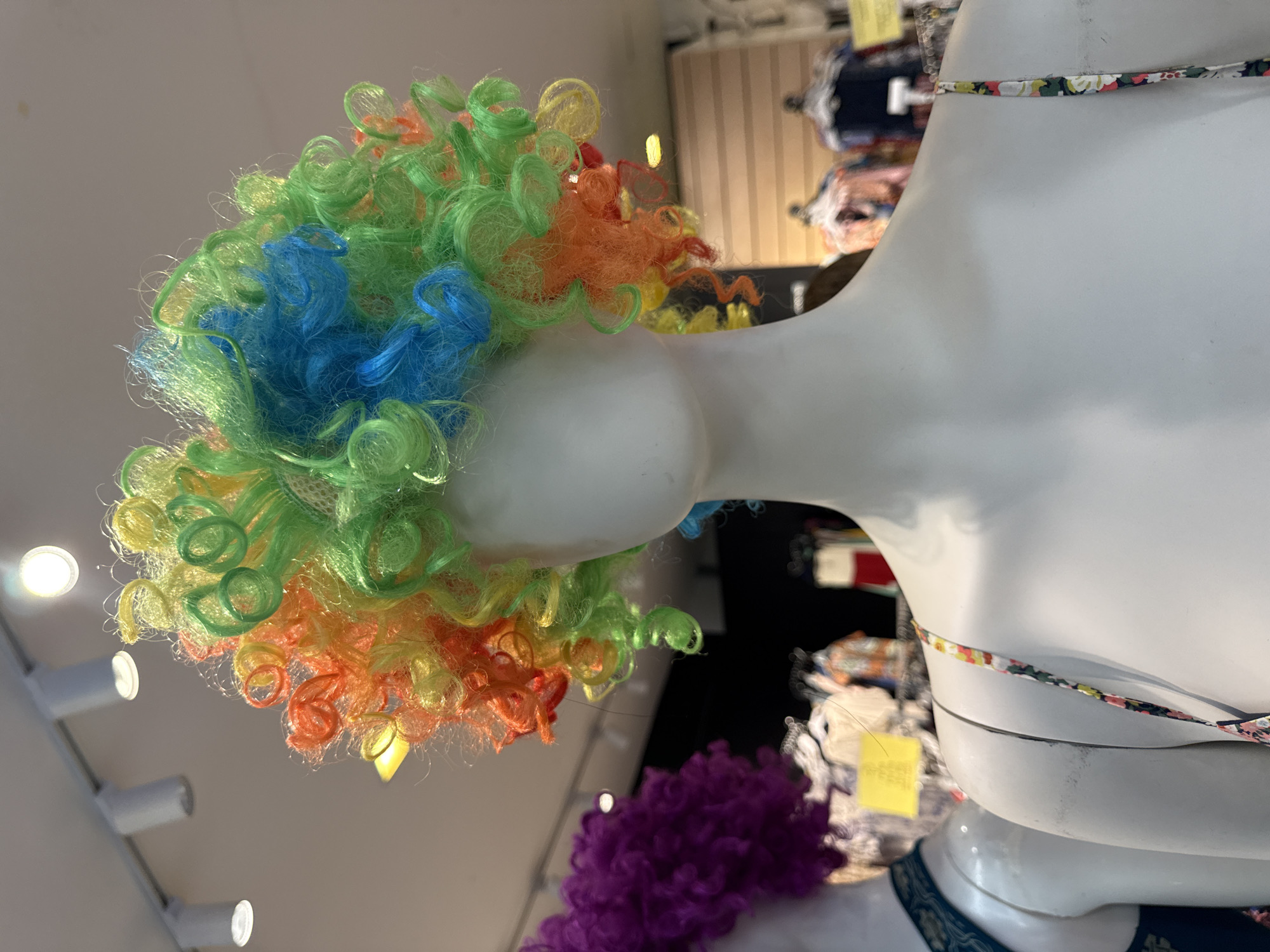 A close-up, eye-level shot shows a white mannequin wearing a brightly colored, curly wig composed of strands of green, blue, orange, and yellow. The wig is styled in a voluminous, chaotic manner, resembling a clown's wig. The mannequin's shoulders and upper chest are visible, and it is wearing a floral-patterned strap. To the left of the main mannequin, another mannequin is partially visible, wearing a purple wig. In the background, the interior of a store is dimly lit, with clothing racks displaying various garments. Overhead, track lighting illuminates the scene. The overall impression is that of a retail environment, likely a costume or novelty shop, with a focus on whimsical accessories. The image was taken in Baan Lamai, Thailand.