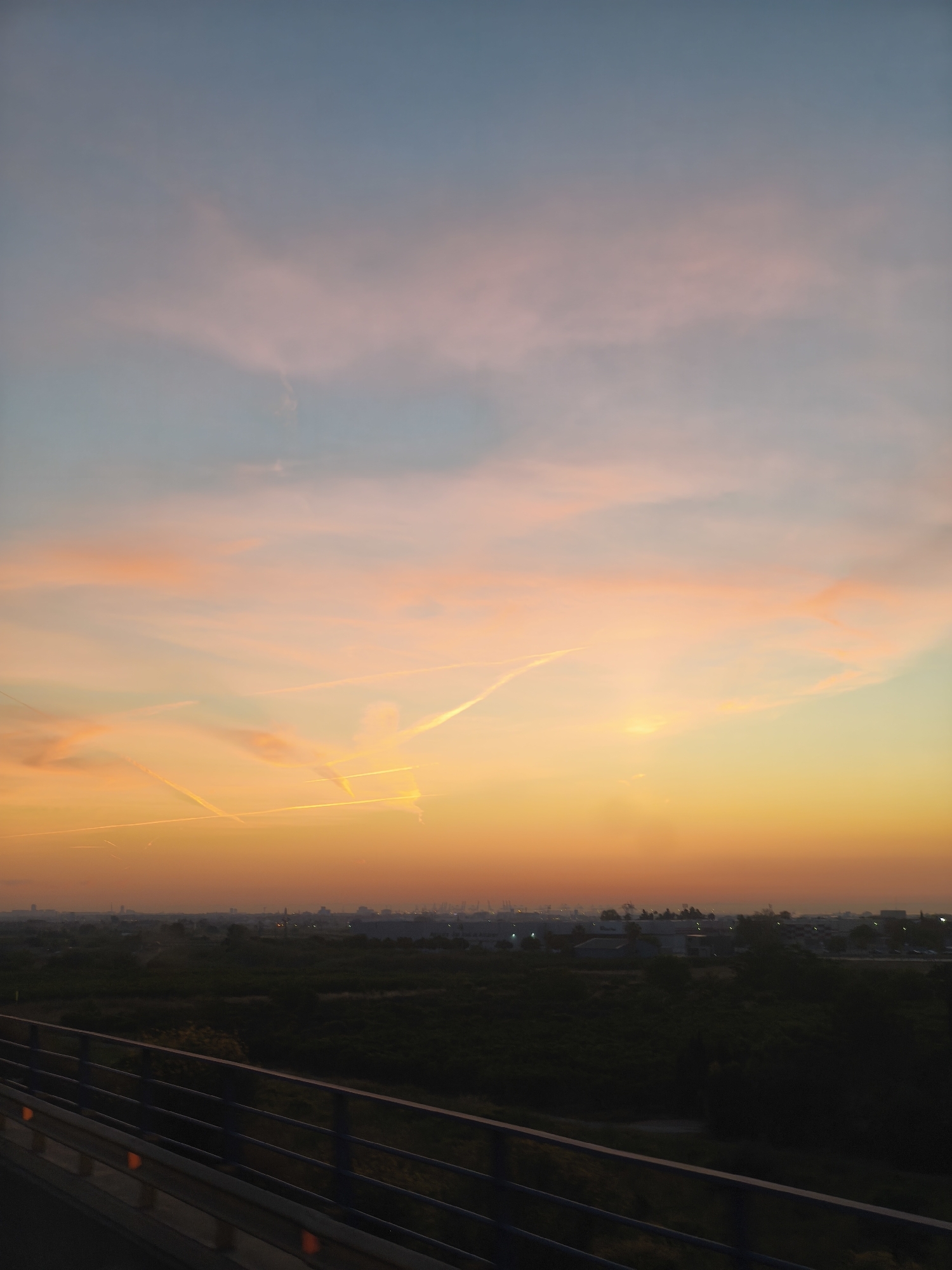 The image shows a view over Alcàsser, Spain at sunset. The sky transitions from blue to orange and pink hues. There are contrails from airplanes. In the foreground is a bridge with metal guardrails. Beyond the bridge, there is dark foliage and vegetation. In the distance, buildings and structures are visible. The image is taken from a high vantage point.