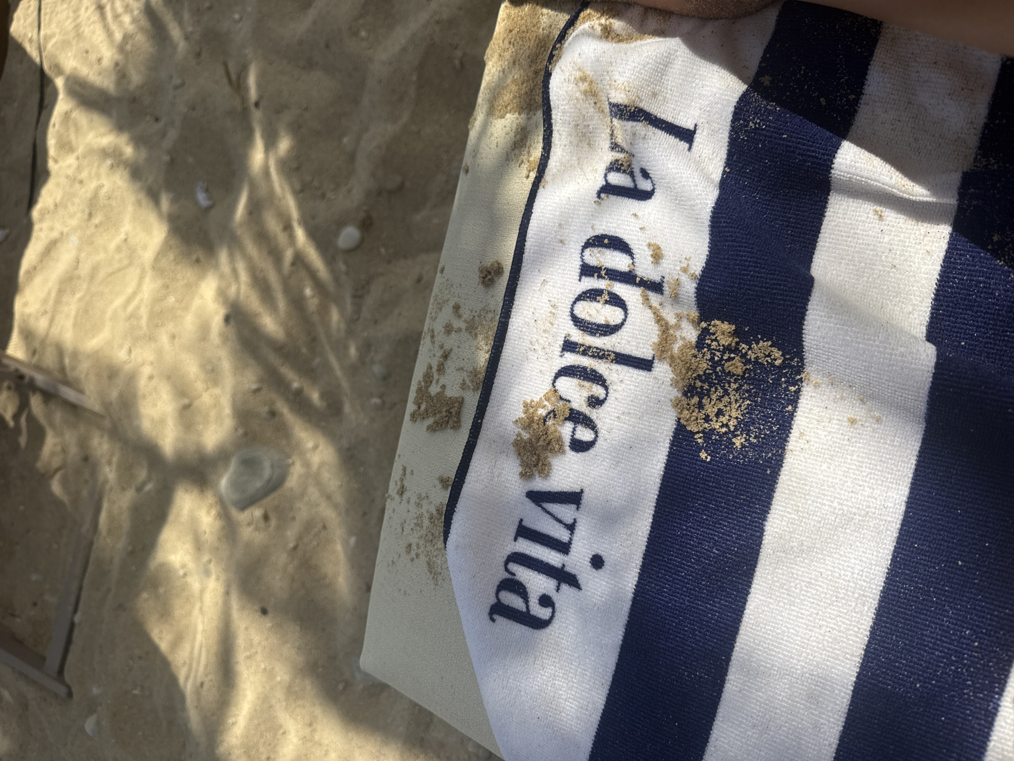 A close-up shot shows a striped towel with the words "La dolce vita" printed on it. The towel is lying on a sandy surface, possibly a beach, and there are small piles of sand scattered across the towel and the surrounding area. The sunlight casts shadows on the sand, creating a dappled effect. The scene appears to be on Ofrynio Beach in Greece.