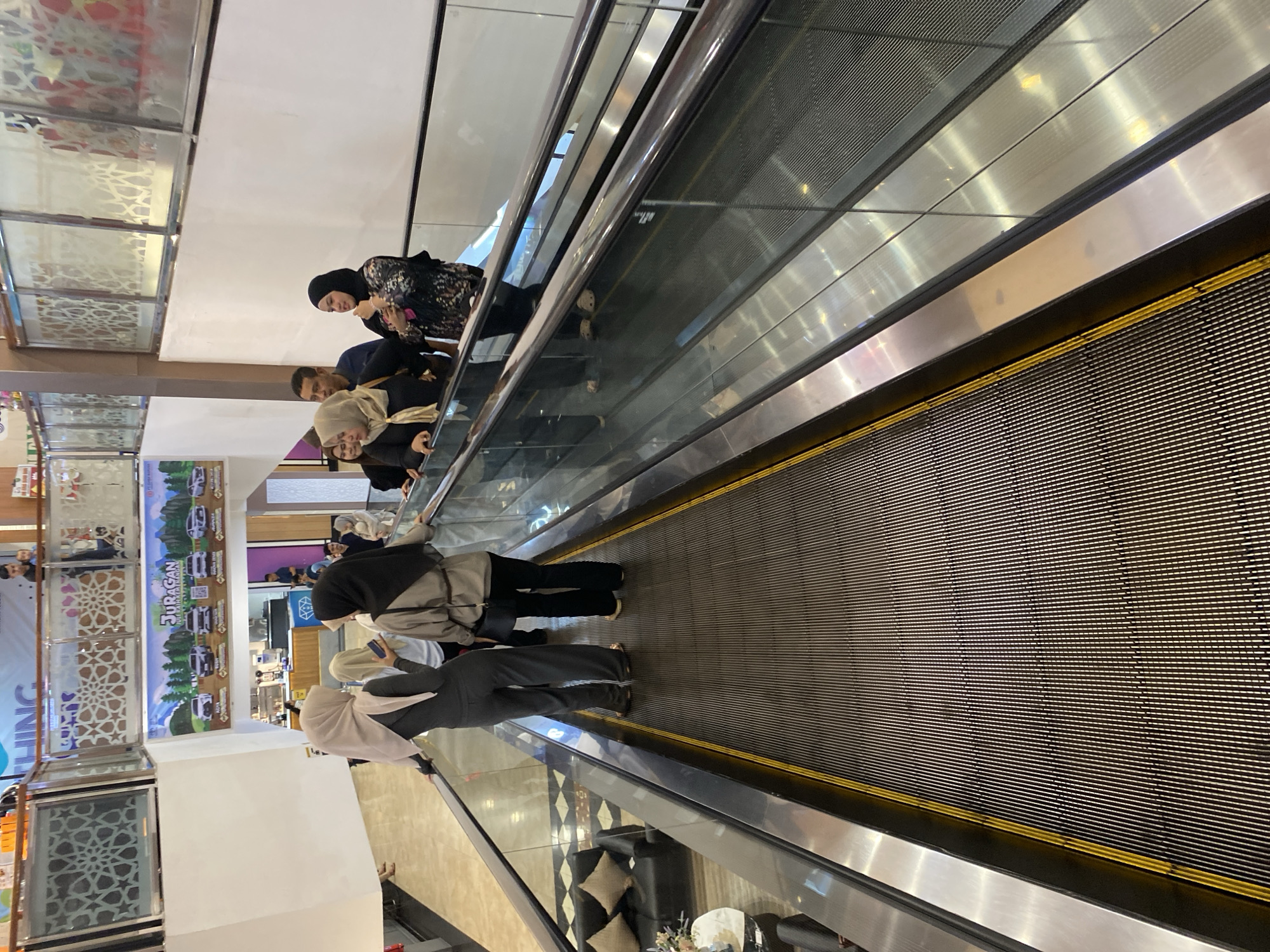 A group of people, predominantly women wearing hijabs, are on a moving walkway inside a brightly lit shopping mall. They are standing and conversing as the walkway carries them forward. In the background, there are advertisements for vehicles and a food court area. The mall features decorative elements on the walls and railings, suggesting a modern commercial environment. This scene is consistent with a typical shopping mall located in Banda Aceh, Indonesia.