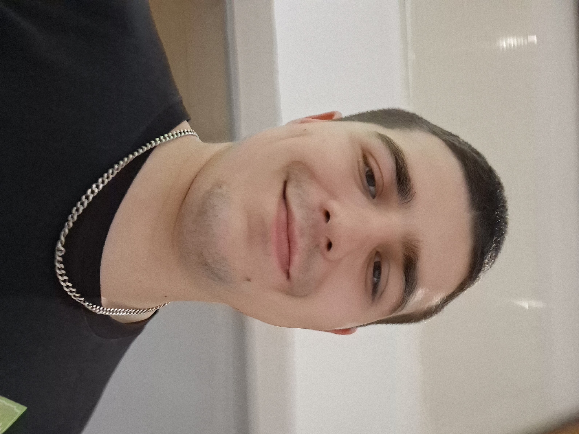 A man is centered in the frame, cropped from the upper chest upwards. He is wearing a black t-shirt and a silver chain necklace. His short dark hair is neatly trimmed. He has fair skin and is smiling gently. The background is a blurry, light-colored wall. The location in Plovdiv, Bulgaria is not evident from the image.