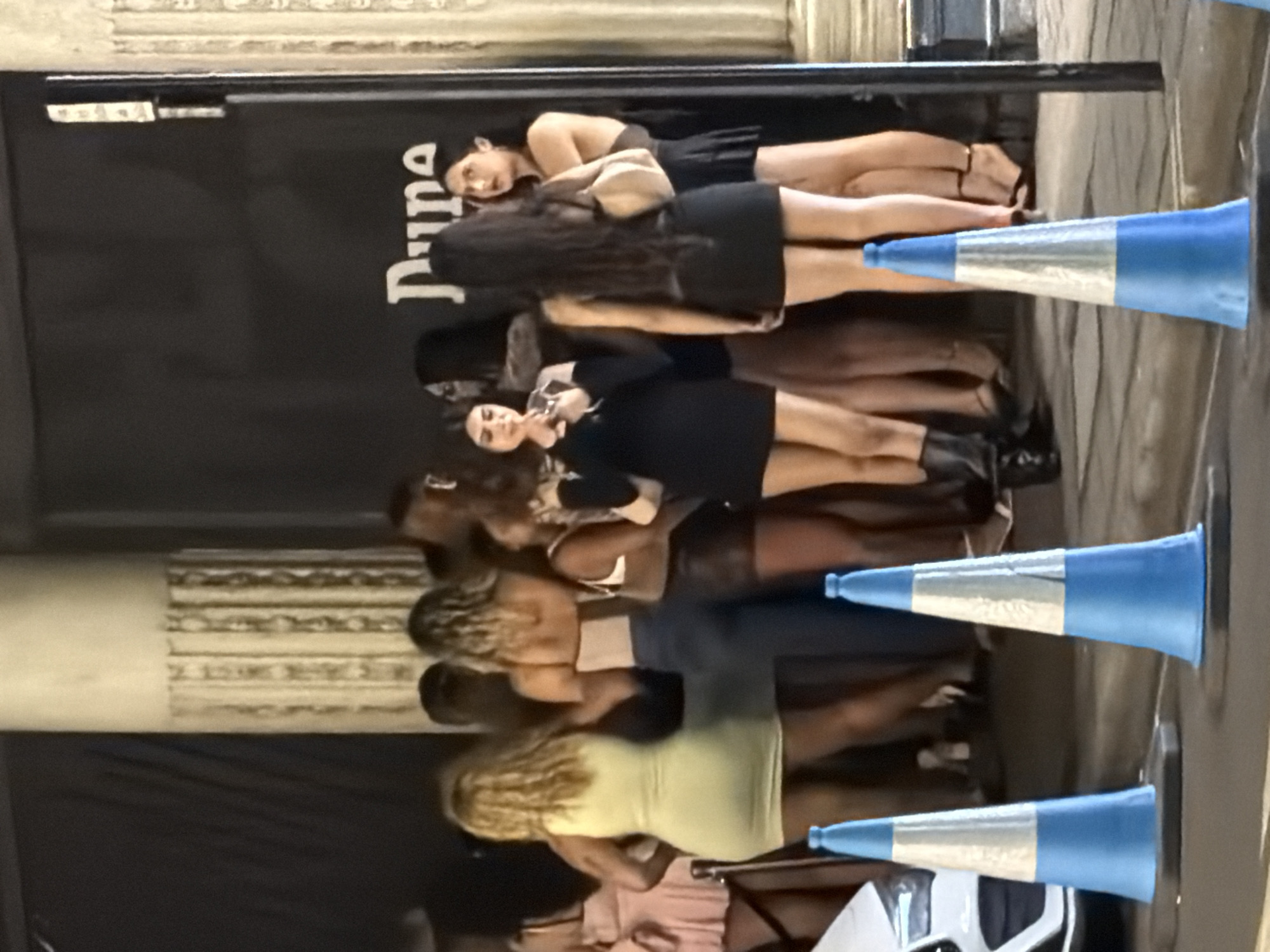 The image depicts a gathering of young women posing in front of a promotion for the film "Dune," likely in London, United Kingdom. The location is suggested by the architectural features, possibly a building exterior with classical columns and moldings, and the overall urban ambiance. The women are dressed in a range of short dresses and skirts, appearing to be attending a promotional event or premiere. Several blue and silver vuvuzelas are present, adding to the celebratory atmosphere. The scene seems to be a photograph or still captured during an event related to the film "Dune."