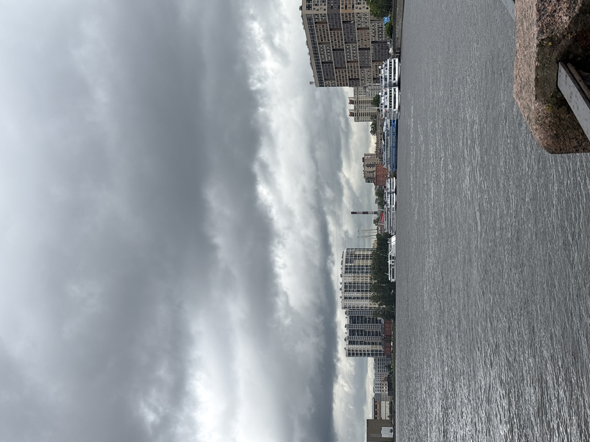 The image shows a view of the Neva River in Saint Petersburg, Russia, under an overcast sky. Multiple river cruise ships are docked along the embankment, with residential and commercial buildings visible in the background. The river surface is textured by wind or currents. Part of the stone embankment is visible in the foreground.