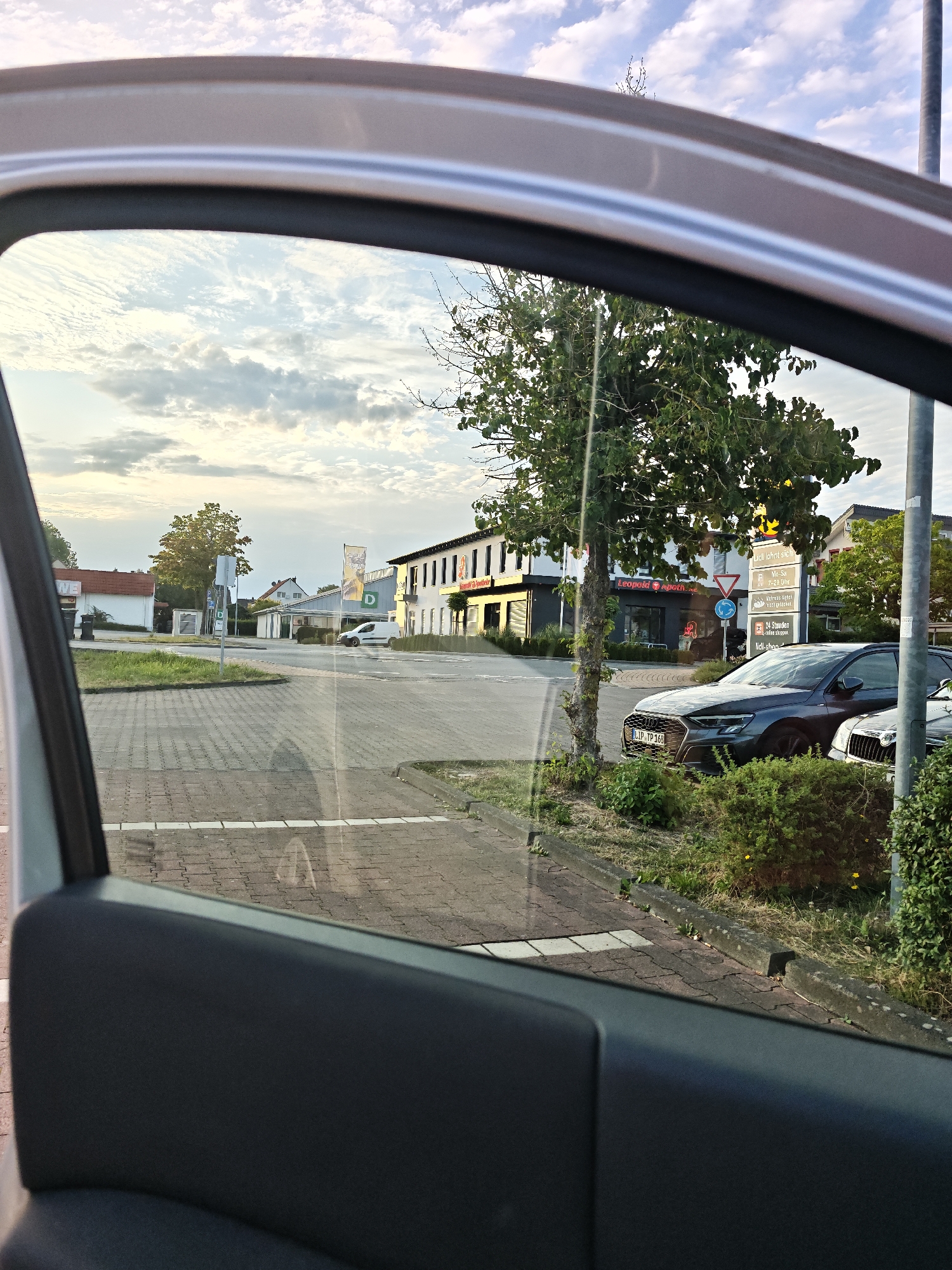 The image captures a view from inside a car, looking out through the open passenger window at a street scene in Leopoldshöhe, Germany. The perspective is from a low angle, focusing on the car's interior door panel in the foreground.
Outside the car, a paved area with interlocking brickwork forms the ground. To the left, a white building with a red roof is visible, with a black trash bin in front. Further down the street, a white van is parked. In the middle ground, several commercial buildings are present. One building is a modern structure with dark facade and illuminated signage for "Leopold Apotheke". Adjacent to it is a "Lidl" supermarket, indicated by prominent yellow and green signage featuring the Lidl logo and listing opening hours. A traffic sign with a circular blue arrow suggesting a roundabout is also visible near the Lidl entrance.
A prominent tree with green foliage stands between the "Leopold Apotheke" and the "Lidl", casting shadows on the pavement. Two cars are parked on the right side of the street: a dark grey Audi in the foreground, and the front of a white car, possibly a Skoda, behind it. The cars are parked near a curb with overgrown green bushes and small yellow flowers. A light pole is also situated to the right of the parked cars.
The sky above is partly cloudy with soft, diffused sunlight, suggesting either early morning or late afternoon. The overall scene depicts a typical suburban commercial area. There are no people visible in the image.