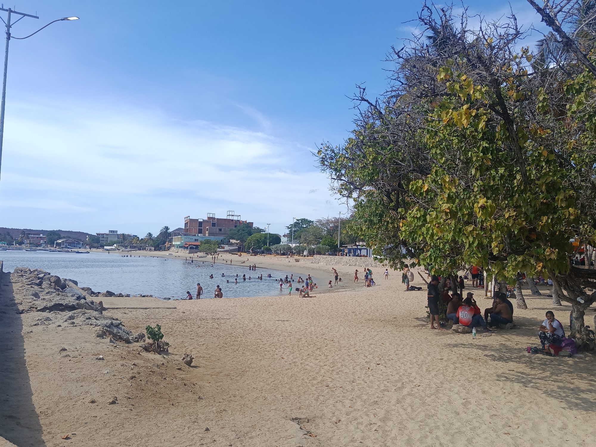 The image shows a beach scene in Juan Griego, Venezuela, with people swimming and relaxing on the sand. In the background are buildings and boats. The beach is sandy, and there are some rocks in the water. A group of people sits under a tree on the right side of the image. The sky is blue with some clouds. A lamppost stands on the left side of the image.