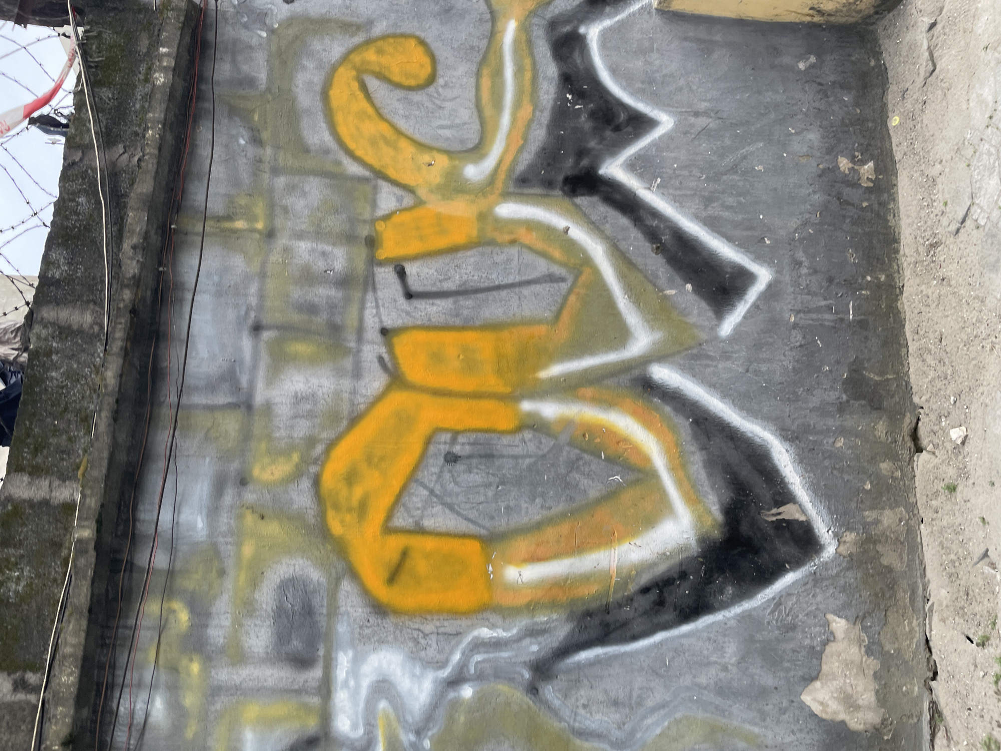 The image displays graffiti on a concrete wall, likely located in Ado, Nigeria. The graffiti features large, stylized letters in bright yellow and white, with black accents. The wall itself is weathered and shows signs of age, with peeling paint and patches of grime. In the background, a concrete ledge with visible wiring and barbed wire suggests an urban or industrial setting. The ground in the foreground is rough and appears to be unpaved or dirt. The overall impression is of an outdoor, possibly derelict, urban space marked by graffiti art. There are no people or specific actions depicted.