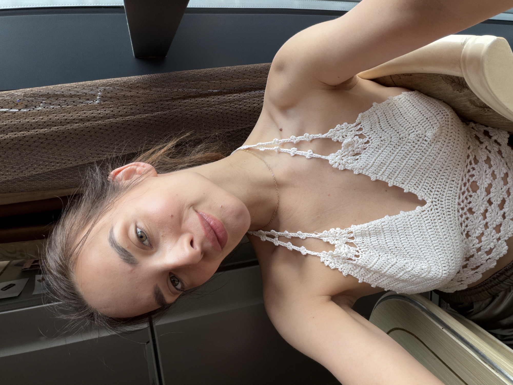 A woman is taking a selfie while sitting in a chair. She is wearing a white crochet halter top and a gold necklace. Her hair is brown and pulled back. She has green eyes and light skin with freckles. Behind her, there is a dark brown curtain and a window. To her left, a portion of a table and what appears to be a refrigerator are visible. The overall environment suggests an indoor setting, possibly an apartment in Ufa, Russia, near a window.