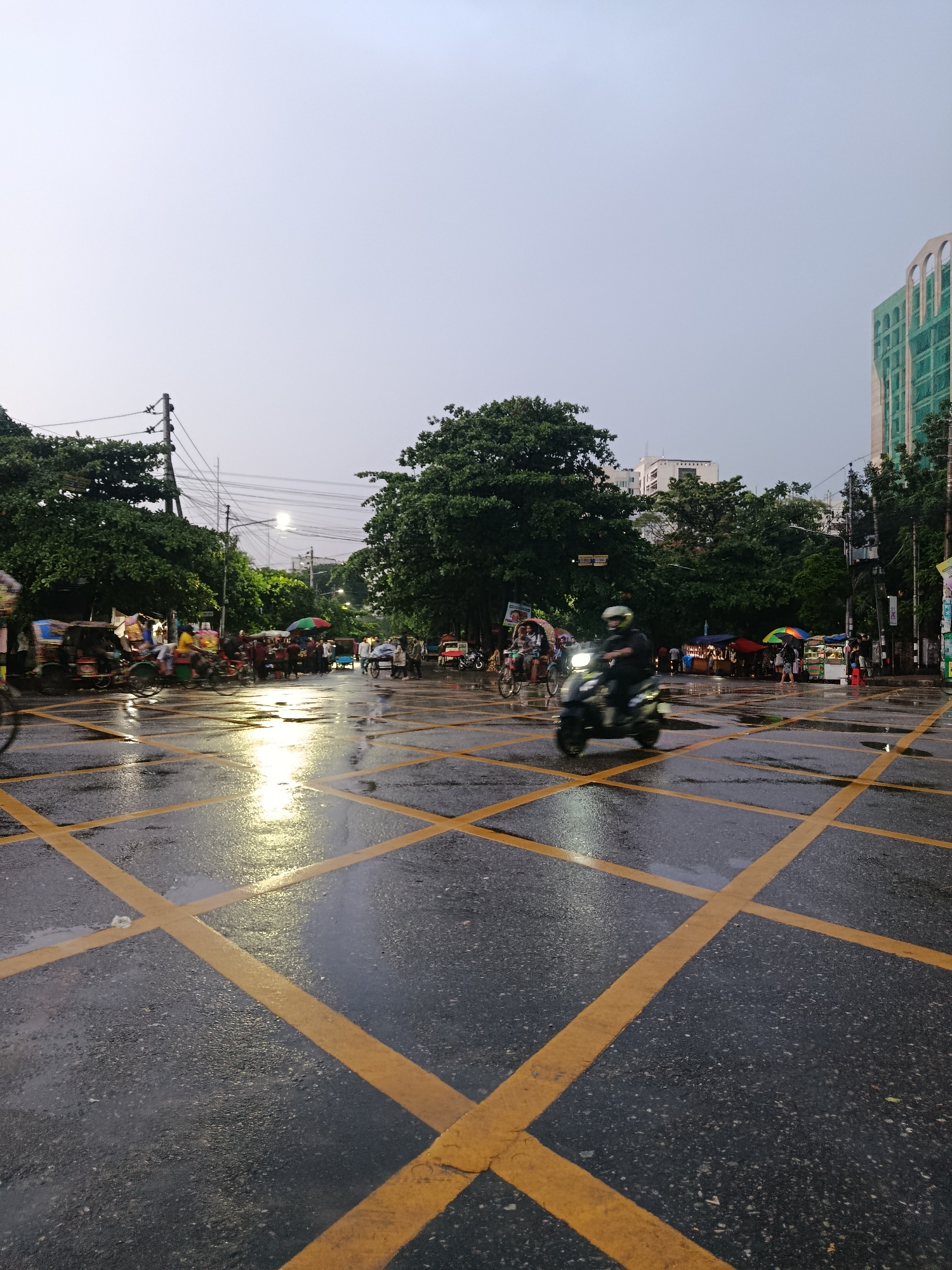 The image shows a wet city street scene in Dhaka, Bangladesh. The road is marked with yellow grid lines, indicating a keep-clear area. Several motorized scooters and bicycle rickshaws are present, some in motion, suggesting active traffic. A few pedestrians are visible near roadside vendor stalls, some shaded by umbrellas. The overall light is dim, likely due to overcast weather or the time of day. Overhead power lines are visible against the sky, and trees line the street. A partially constructed building is visible in the background.