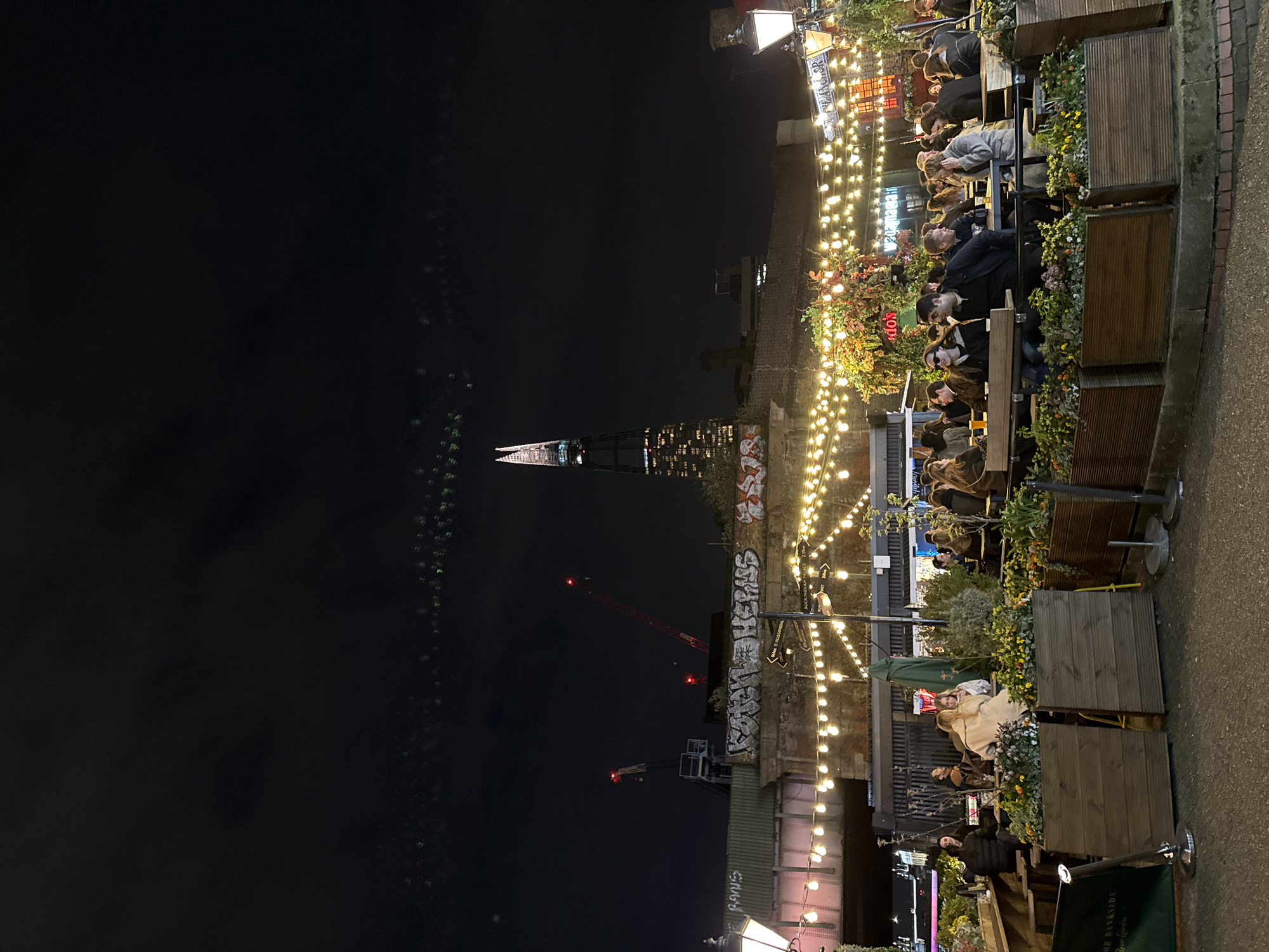 An outdoor dining and social area is depicted at night in London, United Kingdom, specifically near Borough Market under railway arches. The Shard skyscraper is prominently visible in the background, illuminated, with its distinctive spire silhouetted against a dark sky. To the left of The Shard, two red-lit construction cranes are operational.
In the mid-ground, a stone and brick railway viaduct or bridge features extensive graffiti, including the legible phrase "KONCHO HELM". Beneath this structure, an outdoor seating establishment is brightly illuminated by numerous string lights strung horizontally across the scene. Wooden planters, filled with diverse greenery and some flowering plants, define the perimeter of the seating area.
Numerous individuals, estimated between 20-30, are seated at tables within this area. They are primarily adults, casually dressed, engaged in conversation and social interaction. Several patrons have drinks, such as beer, on their tables. A dark green umbrella is visible among the tables.
On the left, a traditional-style street lamp casts a yellowish glow. Another similar lamp is visible on the right. A street sign pole located below the railway arch on the right points towards "St Paul's Cathedral", "BUCKINGHAM PALACE 2.35m", and "BOROUGH MARKET". The ground surface in the immediate foreground is paved, suggesting a pedestrian area.