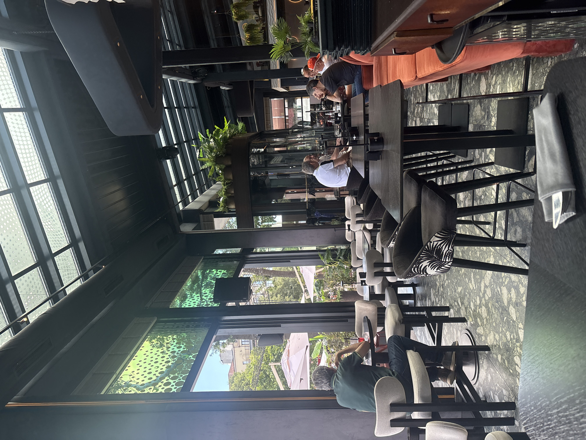 The image shows the interior of a modern restaurant with a high ceiling and a glass roof. Several groups of people are seated at tables, some indoors and some in an outdoor seating area visible through large windows. The interior features dark wood furniture, upholstered chairs and booths, and hanging plants. The floor is tiled with a speckled pattern.
In the foreground, on the left side, a man wearing a green shirt and blue jeans is seated at a round table with a red drink. He is looking out the window. In the background, several other patrons are visible. On the right side of the image, a row of orange booths lines the wall, with a bar area visible beyond.
The overall atmosphere appears to be relaxed and casual, suitable for dining or drinks. The lighting is a mix of natural light coming through the glass roof and windows, and artificial lighting from overhead fixtures. The image is taken from a low-angle perspective, capturing the scale and design of the restaurant.