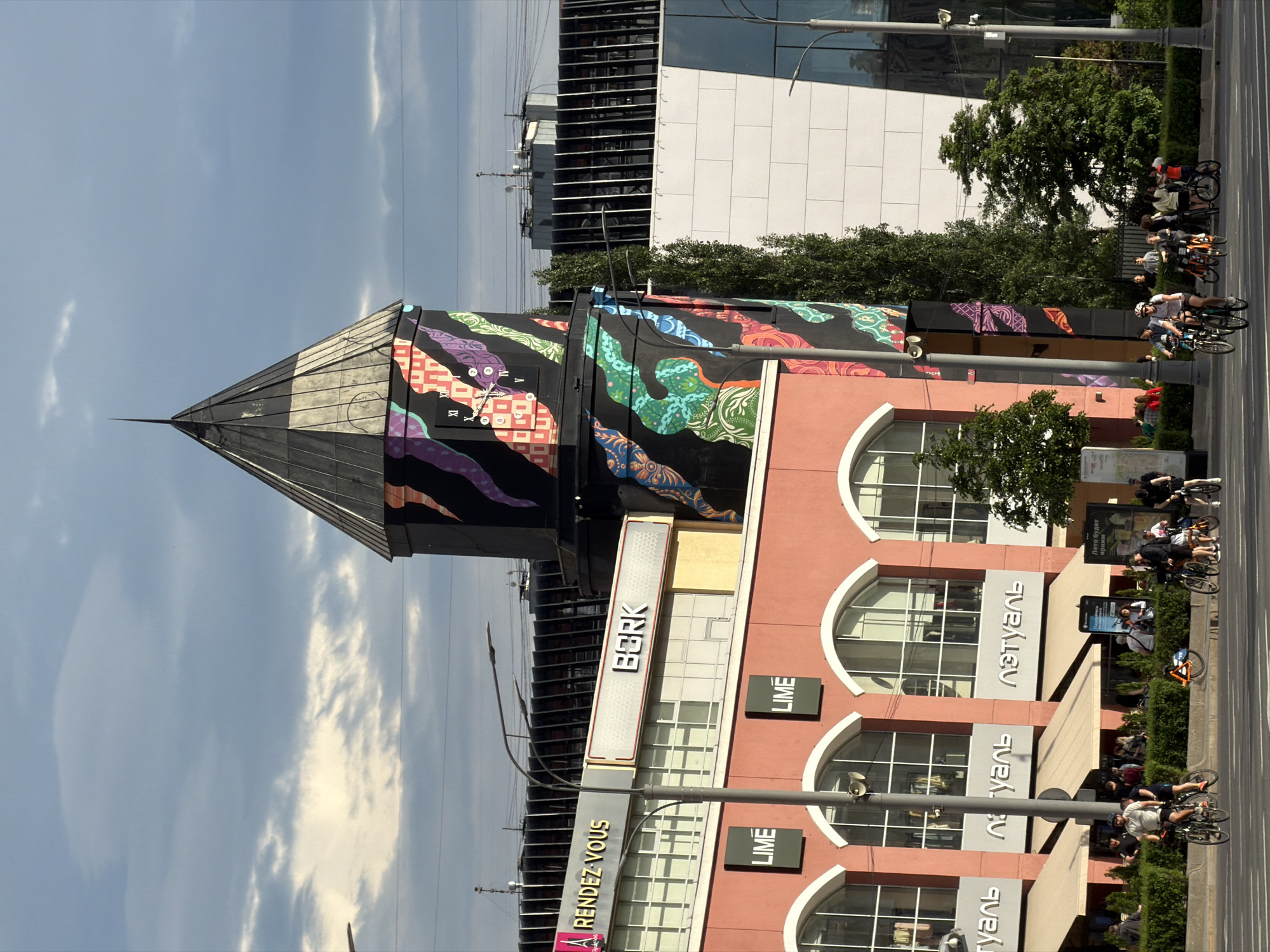 The image depicts a building in Moscow, Russia, featuring distinctive architecture and prominent signage. The building has a pointed, black roof section, with a brightly colored mural with an image of a clock on the side. Below the mural, the building has a sign that says "BORK" in large letters, with signs for "RENDEZ-VOUS" and "LIME" directly below it. The building's façade has large arched windows and the name "туаль" (L'Etoile) vertically displayed. In front of the building, a group of people are riding bicycles along the sidewalk, suggesting a street-level scene. The weather appears sunny, with a clear blue sky. A modern building is visible in the background.