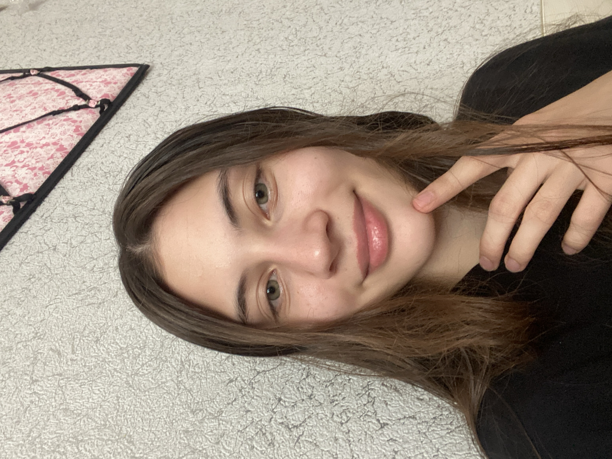 A young woman with long, dark brown hair and green eyes is looking directly at the camera, smiling slightly. She is wearing a black t-shirt. Her right hand is raised to her chin, with her fingers resting on her jawline. The background is a textured gray wall with a pink and white triangular piece of fabric hanging on it. The scene appears to be indoors, likely in a home. There is no specific information to place this image in Votkinsk, Russia.
