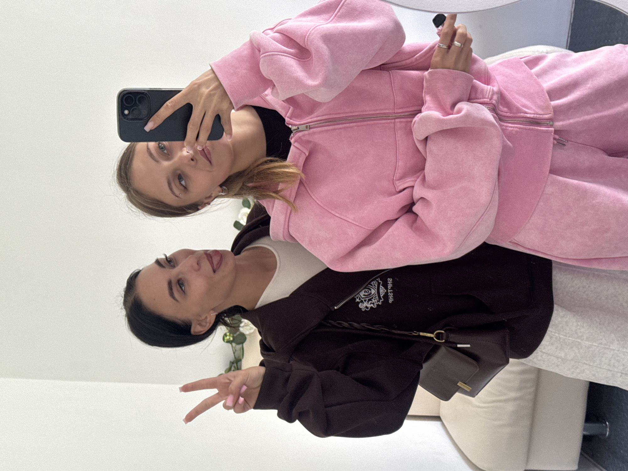 Two young women pose for a selfie in an indoor environment. The woman on the left wears a light pink tracksuit with a full-zip hoodie and matching pants, holding a black smartphone up to take the photo. She has light brown hair and is looking at the camera. The woman on the right wears a dark brown zip-up jacket over a white top, paired with gray sweatpants. She is giving a peace sign with her fingers and sticking her tongue out. The background appears to be a room with light-colored walls and furniture. There is no evidence to suggest the location is in the city of Фоки, Russia.