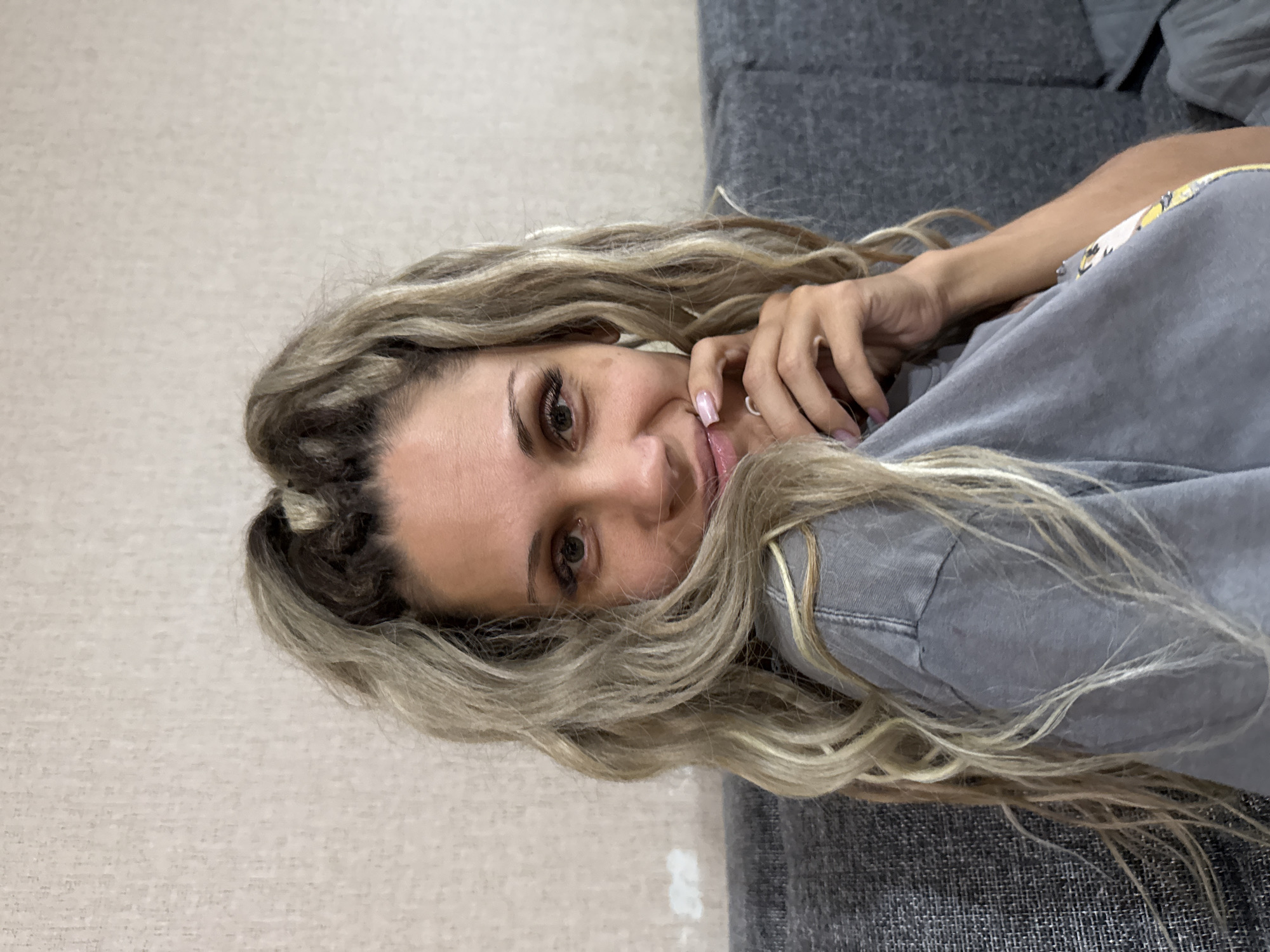 A woman is sitting on a gray couch, leaning her right hand on her chin. Her blonde hair is styled in two pigtails with loose curls. She is wearing a gray t-shirt. The background is a textured, light-colored wall. The scene is indoors, likely in a home, and there is no indication of the city of Rusanovo, Russia, or any urban elements within the image.